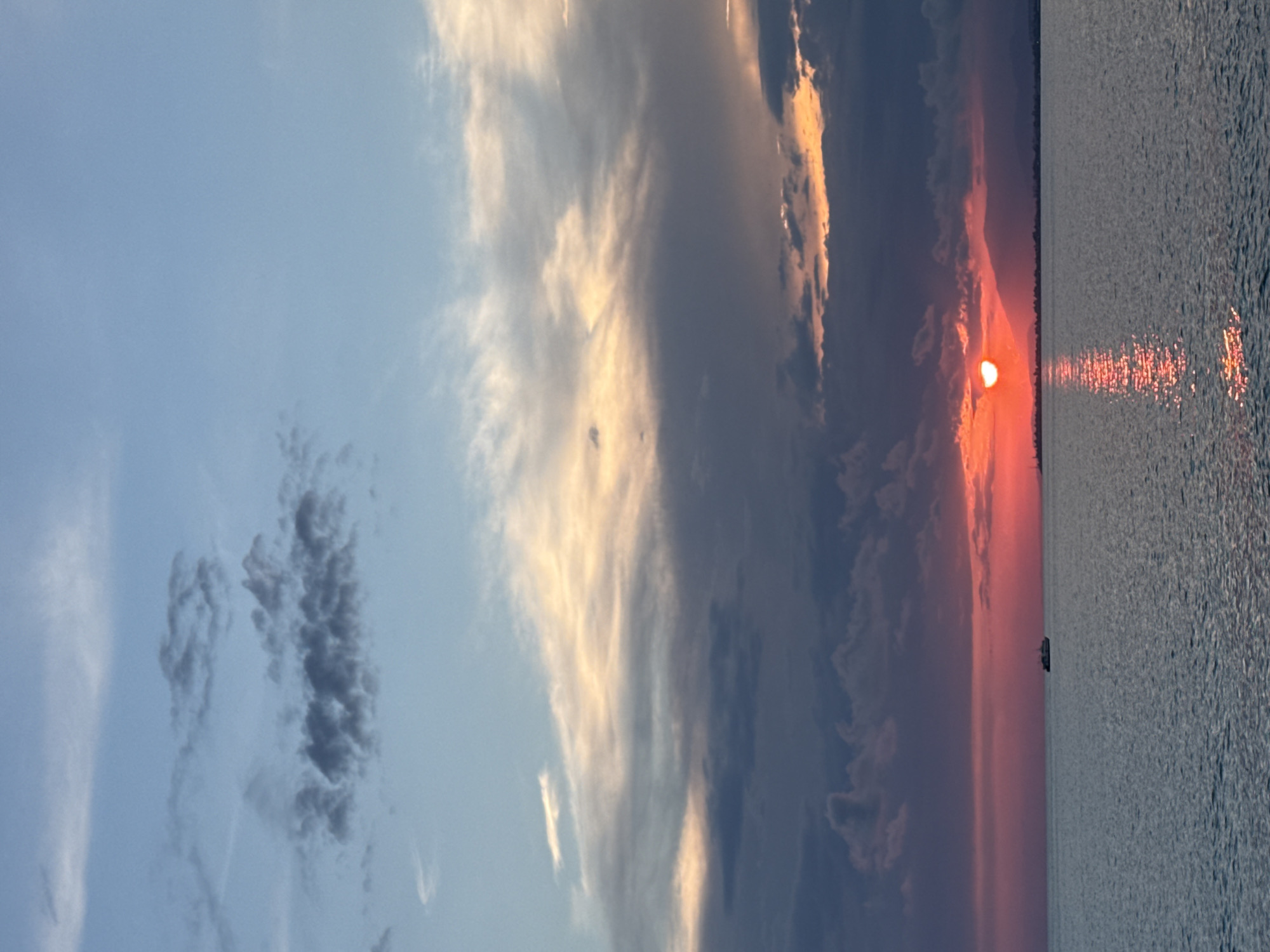 The image captures a serene sunset over the sea in Yuada-Gulrypsh, Abkhazia. The sky displays a dramatic gradient of colors, transitioning from a pale blue at the top to hues of orange, pink, and deep red closer to the horizon. Wispy clouds are scattered across the sky, some tinged with the warm light of the setting sun. The sun itself is a bright, fiery orb partially obscured by clouds, casting a shimmering reflection on the calm water. A lone boat is visible in the distance on the left side of the frame, its silhouette a dark contrast against the vibrant sky. The horizon line is marked by the dark outline of distant mountains and a faint indication of a shoreline. The water is a deep, muted blue, rippled by gentle waves.