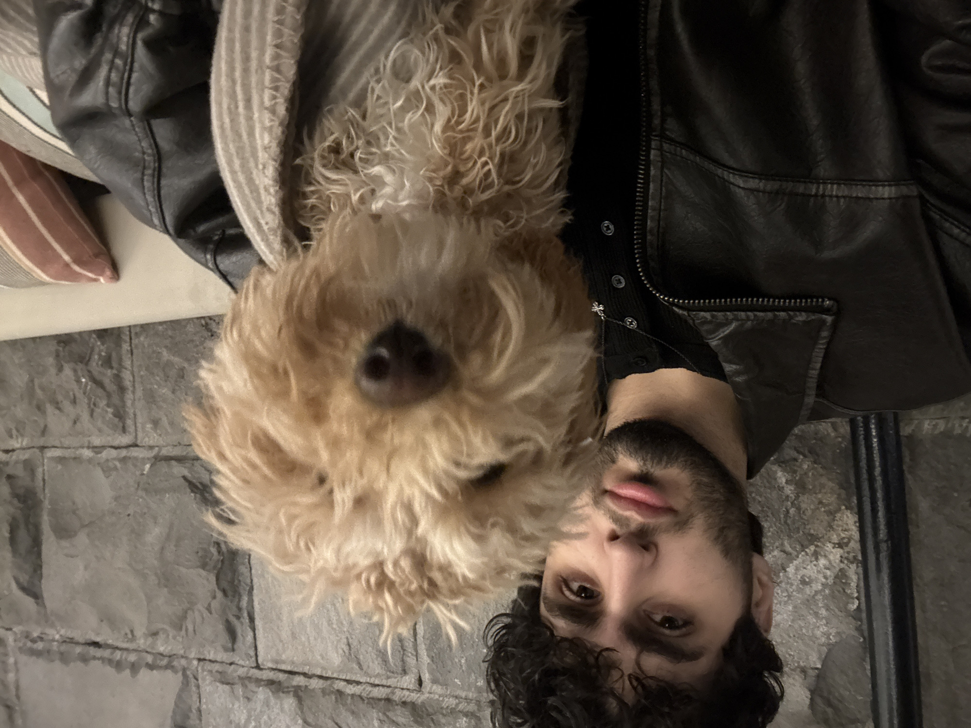 A man with dark curly hair and a beard poses with a golden doodle dog. The man is wearing a black leather jacket over a button-down shirt with a small cross necklace. They appear to be on a stone or brick floor. It's unclear if the location is Yerevan, Armenia.