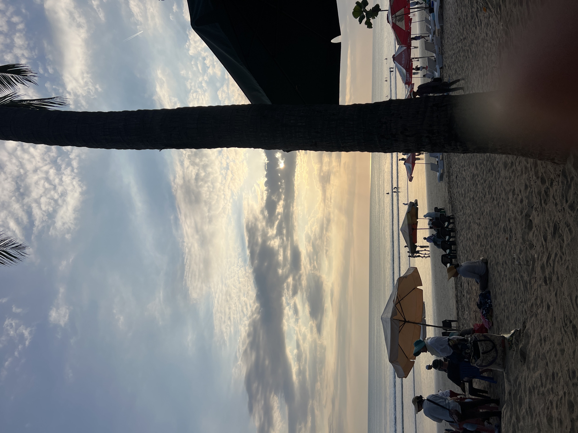 The image depicts a serene beach scene in Kuta, Indonesia, during what appears to be sunset or sunrise. The sky is a blend of pale blue and soft yellow, with wispy clouds illuminated by the low-hanging sun, casting a warm glow on the horizon. The ocean is calm, with gentle waves lapping at the shore.
In the foreground, a tall palm tree dominates the right side of the frame, its dark silhouette contrasting with the bright sky. A large, dark umbrella is also visible, casting a shadow over a portion of the sand. The beach is populated with several beachgoers. Some individuals are seen standing, while others are seated or walking. Beach umbrellas, some colorful and others a neutral beige, are dotted across the sand, providing shade for loungers and small groups of people. Several beach chairs are arranged neatly under some of the umbrellas.
In the mid-ground, the ocean is dotted with several figures, some appearing to be on surfboards, indicating water activities. The overall atmosphere is peaceful and relaxing, typical of a tropical beach destination. The sand itself is a light brown, with footprints and slight undulations indicating its texture. The image captures a moment of tranquility, with the natural beauty of the beach and the gentle activity of its visitors contributing to a sense of calm.