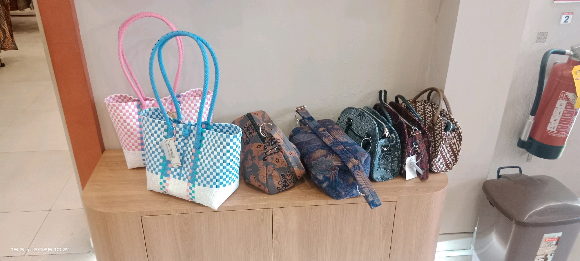 An indoor medium shot captures a collection of woven and fabric bags displayed on a light-colored wooden cabinet. The bags are arranged from left to right. The two leftmost bags are pink and blue, made of woven plastic with white trim and blue handles. The blue bag has a white tag attached to its handle. To the right of these are several fabric bags with batik-style patterns. These bags vary in color, including shades of brown, dark blue, and grey, with some featuring intricate floral or geometric designs. Most of these fabric bags have zippers and metal ring details. In the background, a portion of a red fire extinguisher is visible mounted on a light-colored wall, along with a sign with the number '2' on it. To the left of the bags, a hint of a display of fabric or clothing is present. The bottom left corner of the image contains a timestamp indicating "15 Sep 2026 10.21". The overall environment suggests a retail setting or a display area within a shop in Surakarta, Indonesia, possibly a souvenir or craft store given the presence of batik-patterned items.