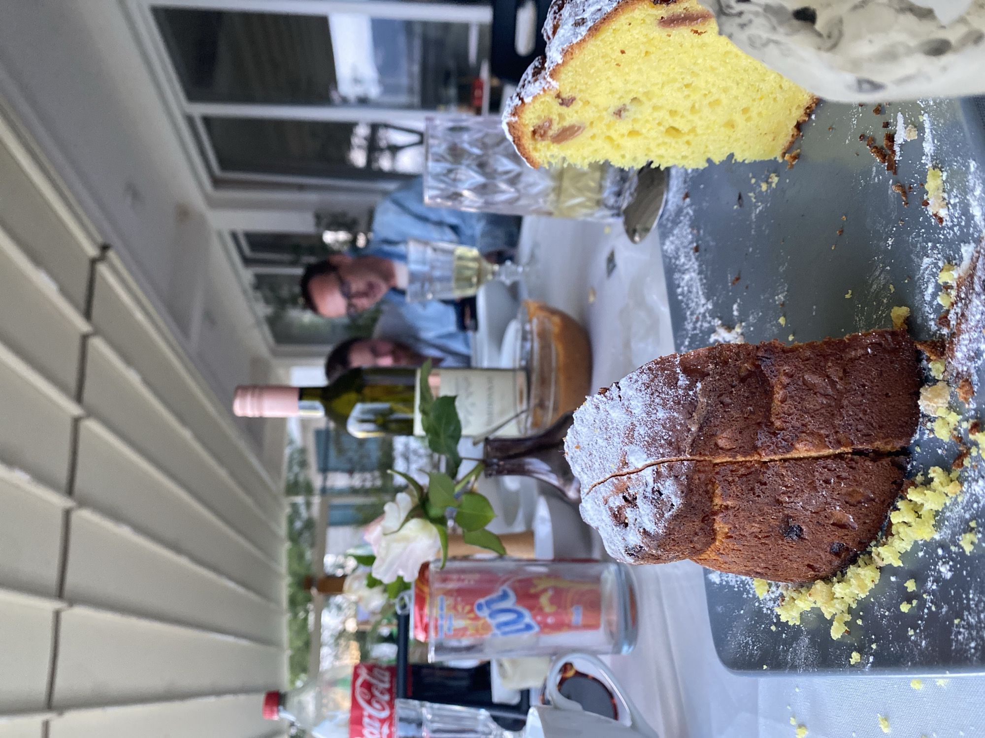 The scene depicts a partially set table with two types of cakes, wine, and soft drinks. One cake is yellow, dusted with sugar, and appears to contain fruit. The other is a darker loaf cake, also dusted with sugar and showing signs of being cut. Beverages include wine in bottles with one having a pink neck seal, and a bottle of Coca-Cola and a bottle of juice. A vase with white flowers is present. In the background, two people are visible seated at the table, one wearing glasses and a blue shirt. The environment suggests an outdoor or semi-outdoor setting, possibly a patio, with a window or opening visible in the background. The city of Trebesin, Montenegro, is not discernible from the given information.
