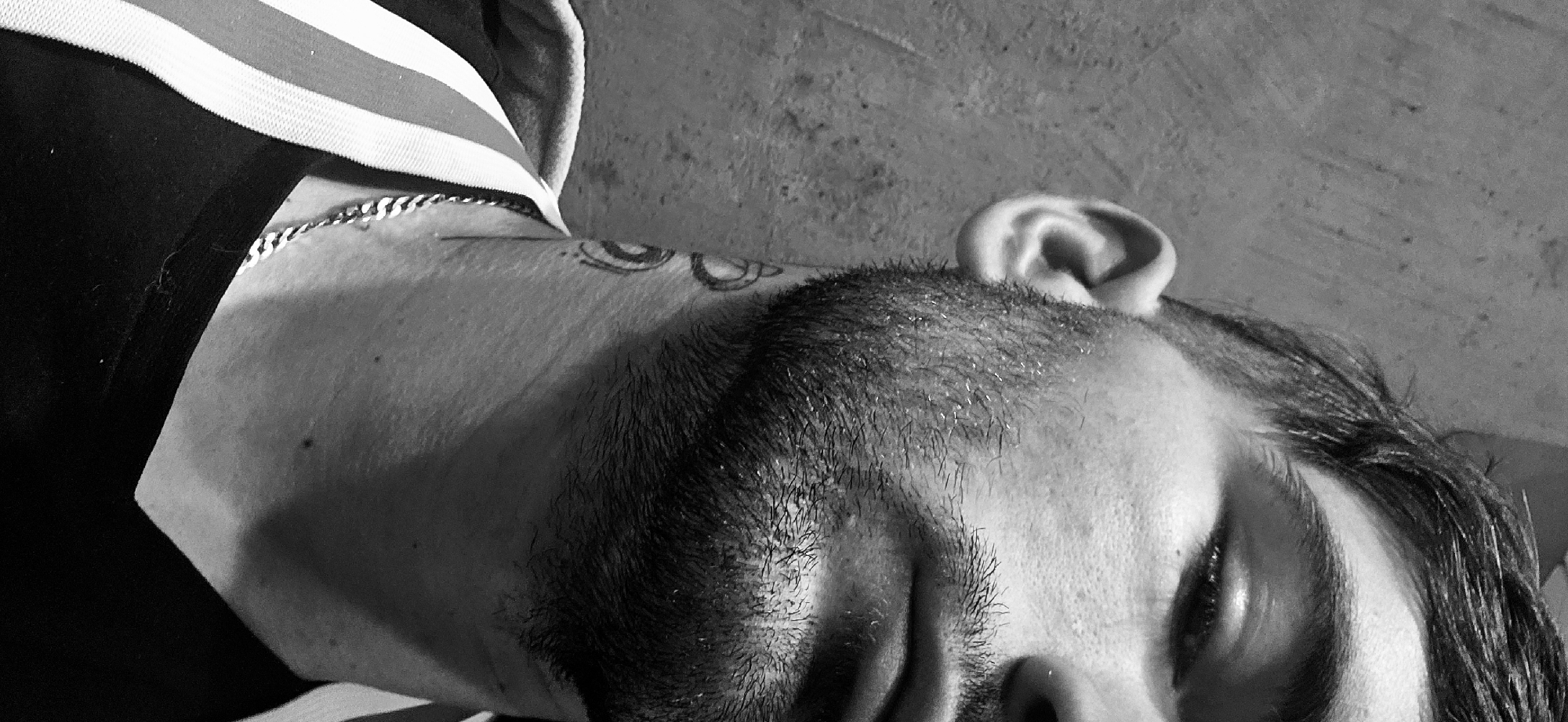 A black-and-white photograph captures a close-up, low-angle view of a man lying down. He is seen from the top of his head, with his face partially visible. His eyes are closed. He has short, dark hair on top, with longer hair along the side of his head. His ear is prominent near the top of the frame. On his neck, just below the hairline, is a tattoo. He is wearing a dark shirt with white stripes on the shoulder. A silver chain is visible around his neck. The background is a textured, solid surface. There is no clear evidence to establish the location as Plovdiv, Bulgaria.