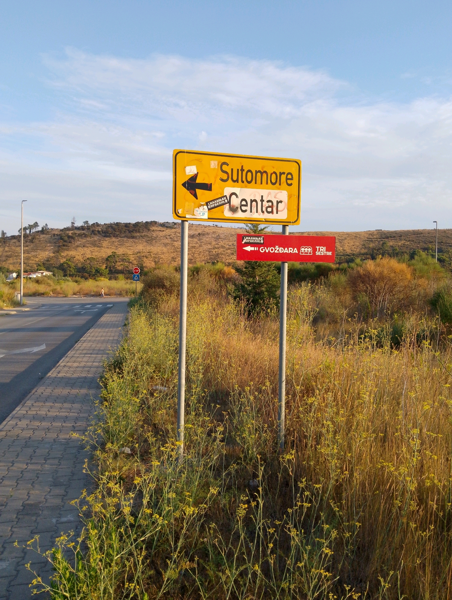 A yellow directional signpost indicating "Sutomore Centar" with an arrow pointing left is positioned next to a paved walkway and a road in a semi-rural setting. The sign is partially covered with stickers. Below this sign, a smaller red sign points left and displays the text "Gvoždara" and "Tri Sestre" with accompanying graphics. The surrounding environment is characterized by dry, tall grass and sparse bushes. In the background, a gentle, treeless hill rises, with a few scattered trees and buildings visible on its lower slopes. A street light stands on the left side of the road, and a stop sign with a pedestrian crossing symbol is present further down the road. The sky is clear with some wispy clouds.