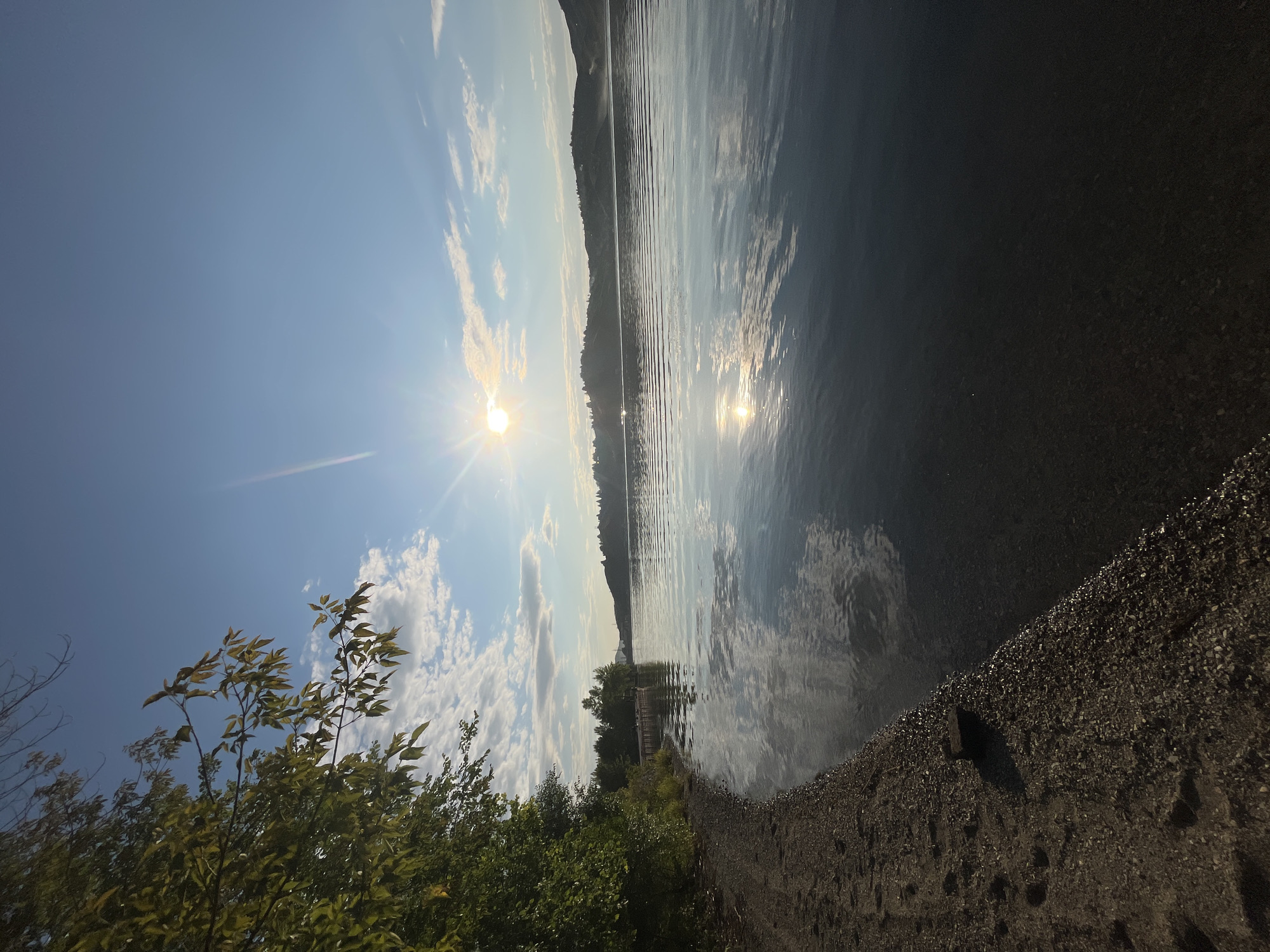 A serene natural landscape unfolds by a calm body of water. The foreground is dominated by a sandy and pebbly shoreline, with a few dark rocks scattered on the ground. Lush green trees, with sunlight filtering through their leaves, line the left side of the frame. The water's surface is remarkably still, reflecting the clear blue sky, scattered white clouds, and the bright sun with a distinct lens flare. In the distance, rolling hills with scattered vegetation form the horizon. A small wooden pier extends into the water from the left bank. The overall scene evokes a sense of tranquility and natural beauty, likely captured in the early morning or late afternoon due to the low angle of the sun. Without further information, the specific location in Oskemen, Kazakhstan, cannot be identified from the image alone.