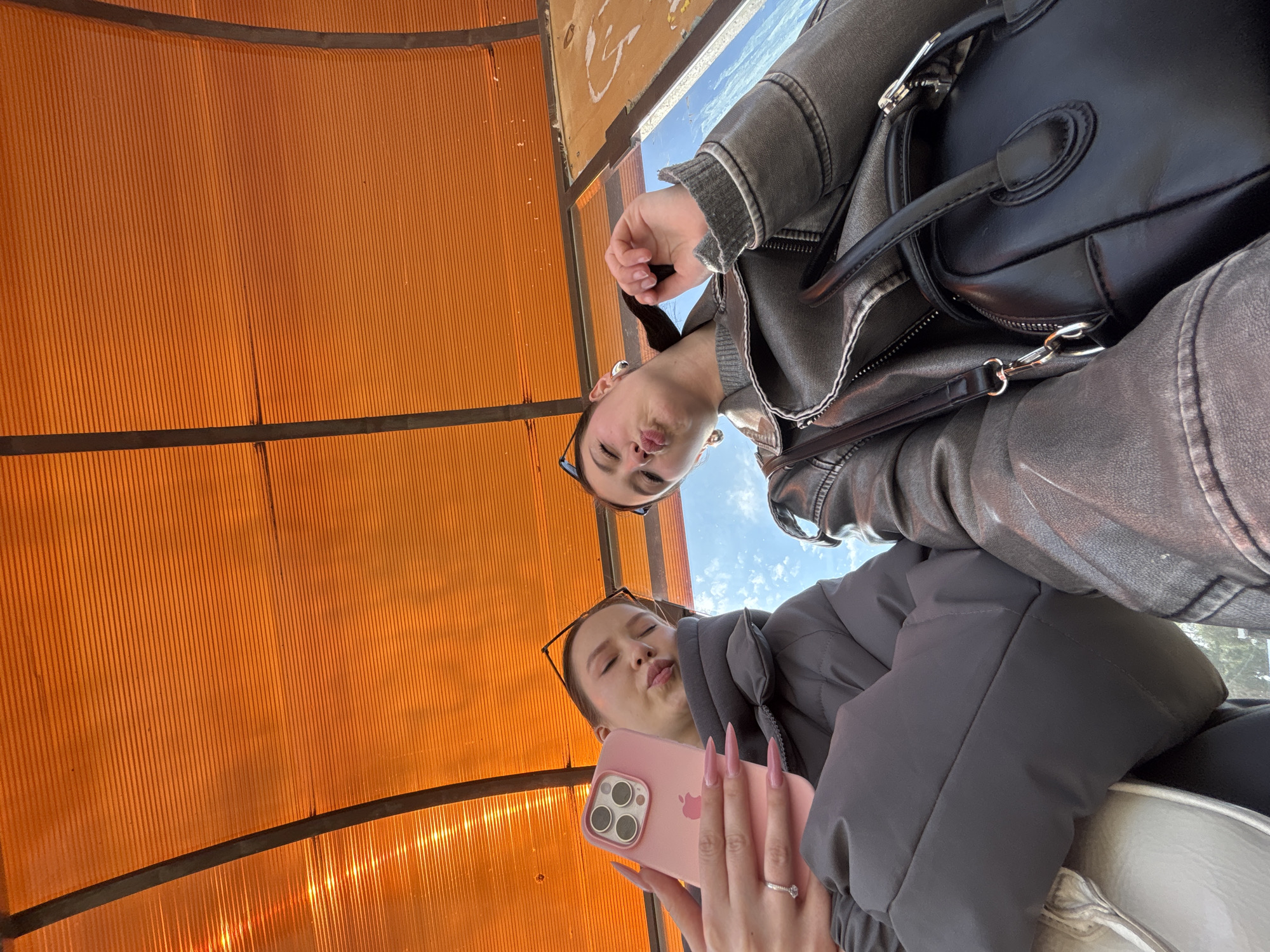 The image, rotated 90 degrees clockwise, presents a selfie of two young women captured from a low angle. Both subjects are looking towards the camera with playful "duck face" expressions, featuring pursed lips.
The woman positioned closer to the camera, on the left side of the frame (when viewed upright), wears a dark grey puffer jacket. She is holding a pink iPhone, which has three visible camera lenses, indicating she is taking the photograph. Her long, pointed nails are painted a matching pink, and she wears a ring on one finger. Sunglasses are pushed up onto her head.
The second woman, further back and to the right, is dressed in a dark grey or brownish leather-look jacket over a grey ribbed sweater. She sports round earrings and also has sunglasses resting on her head. A black handbag with silver hardware is visible on her shoulder or arm.
The background primarily consists of a translucent, corrugated orange or amber roof structure, likely part of a covered outdoor area such as a bus stop, shelter, or walkway in Chaikovsky, Russia. Dark support beams segment this structure. Through an opening or window in the upper right, a clear blue sky with a few white clouds is visible, suggesting the photo was taken during daylight hours. Reflections of sunlight create bright vertical streaks on the ribbed orange material in parts of the frame. No discernible text is clearly visible, though there are some faint, illegible markings on the orange corrugated surface. The overall mood is casual and lighthearted.