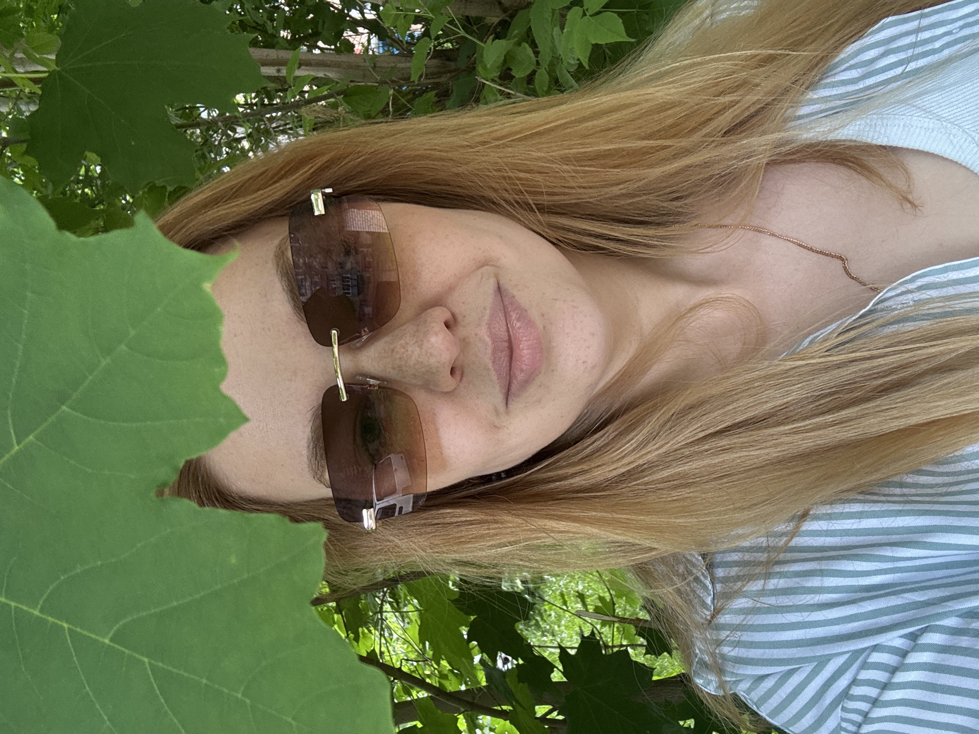 This image, rotated 90 degrees counter-clockwise, depicts a person, likely a woman, posing outdoors amidst abundant green foliage in Rusanovo, Russia. The individual has long, light blonde to golden-brown hair that falls around their face and shoulders. They are wearing large, rectangular brown-tinted sunglasses with gold-colored metal accents, which obscure their eyes. A subtle, soft smile is visible on their lips, and light freckles dot their nose and cheeks. Reflections in the sunglasses show hints of light-colored buildings or structures and the sky, indicating an urban or suburban outdoor environment.
The person is dressed in a light blue and white striped shirt, and a delicate, thin gold-colored chain necklace is visible on their chest, peeking through their hair. Large, vibrant green leaves, possibly from a maple tree, frame the left side of the person's face in the foreground and also form a dense, leafy background. The bright, natural light suggests it is daytime, likely a sunny or partly cloudy day, creating a fresh and serene atmosphere. The overall mood is relaxed and calm. No visible text is present in the image.