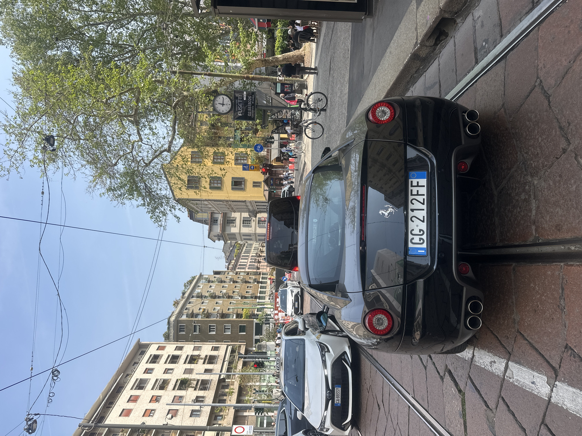 The image depicts a street scene in Milan, Italy. In the foreground, a black Ferrari is parked on a street paved with brick and tram tracks. The license plate reads "GG-212FF". Behind the Ferrari, a white car is also parked. Other cars and vehicles are visible further down the street, along with pedestrians walking on the sidewalks.
In the background, there are buildings of varying heights and architectural styles. A clock is visible on a building, and a sign with a phone number is nearby. Overhead, there are power lines stretching across the street. A tree with green leaves is also visible. The sky is clear and blue.