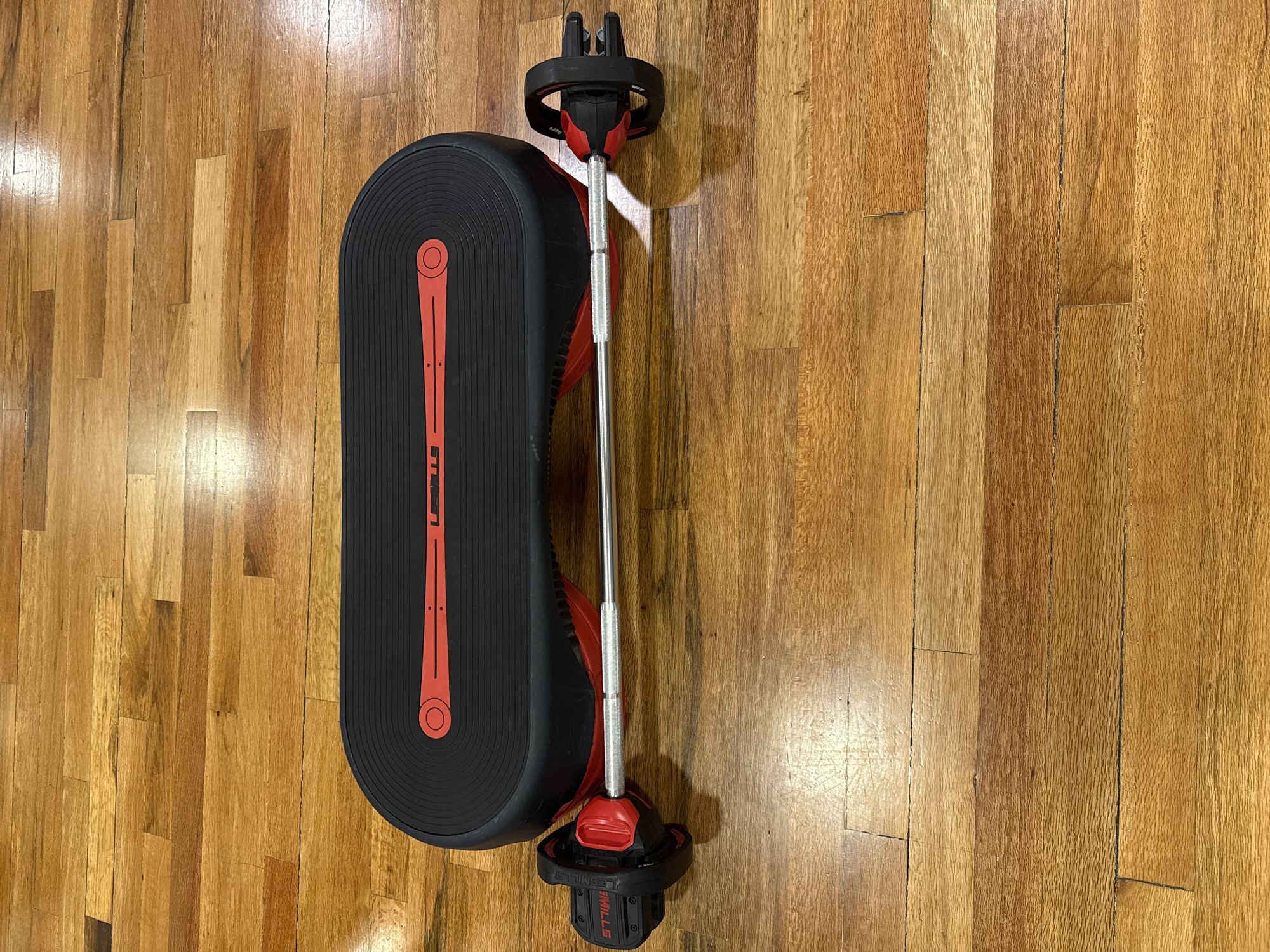 The image shows a black Les Mills Smartstep platform with red accents and a chrome Les Mills Smartbar with red attachments lying on a wooden floor. The floor consists of parallel planks of wood with varying shades of brown. There is no evidence of the city of Puebla, Mexico, or any people, subjects, actions, or interactions in the image.