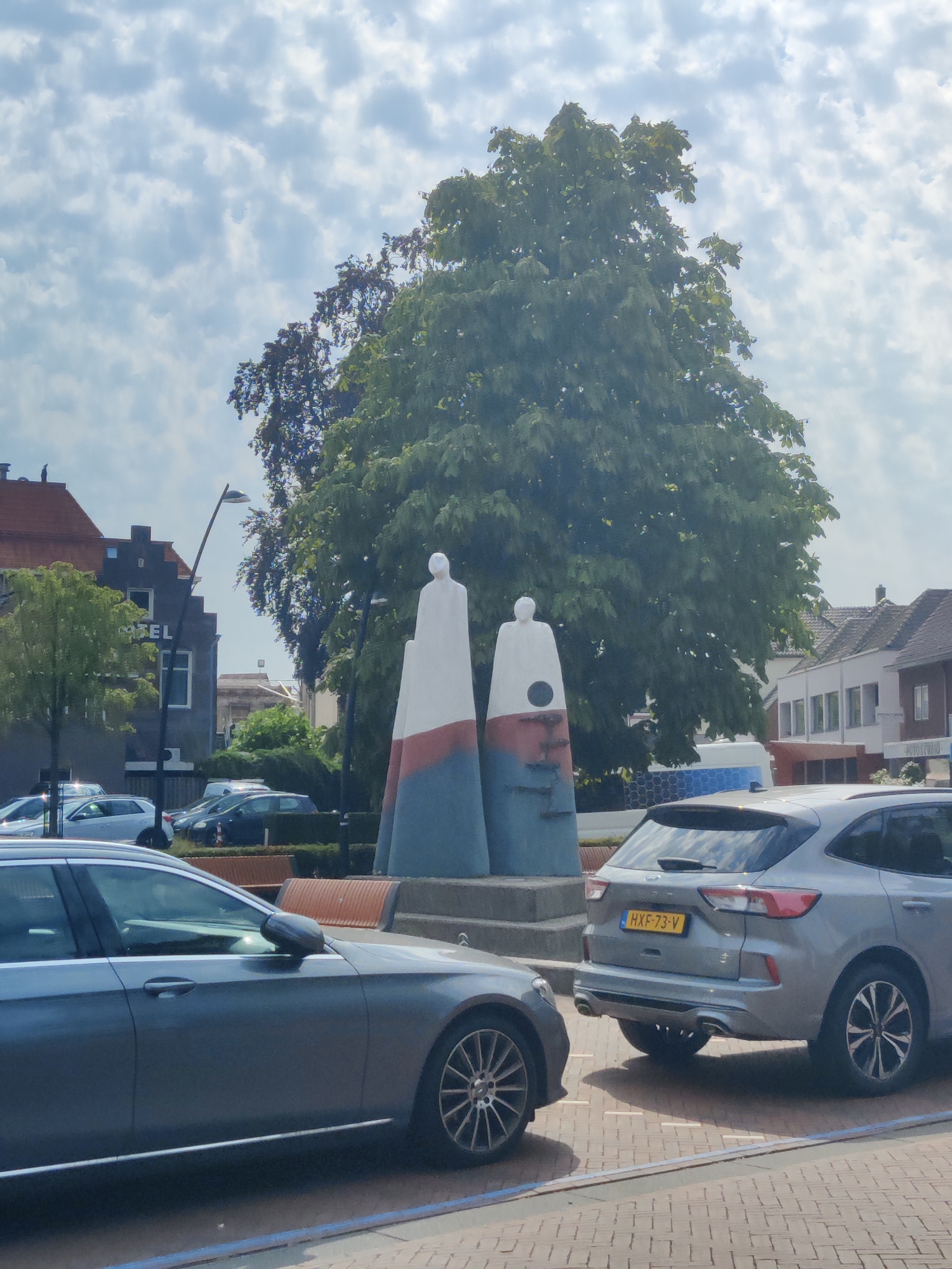 The image depicts a street scene in Winterswijk, Netherlands, featuring a public sculpture of two stylized figures. The sculpture, composed of three conical sections in white, red, and blue, stands centrally in the frame. In the foreground, two cars are visible: a grey sedan on the left and a silver SUV on the right. The SUV has a license plate reading "HXF-73-V". Behind the sculpture, a large green tree dominates the background, with a few buildings peeking through its foliage. The sky is partly cloudy. The area appears to be a public square or street with a paved surface and a bench visible near the sculpture.
