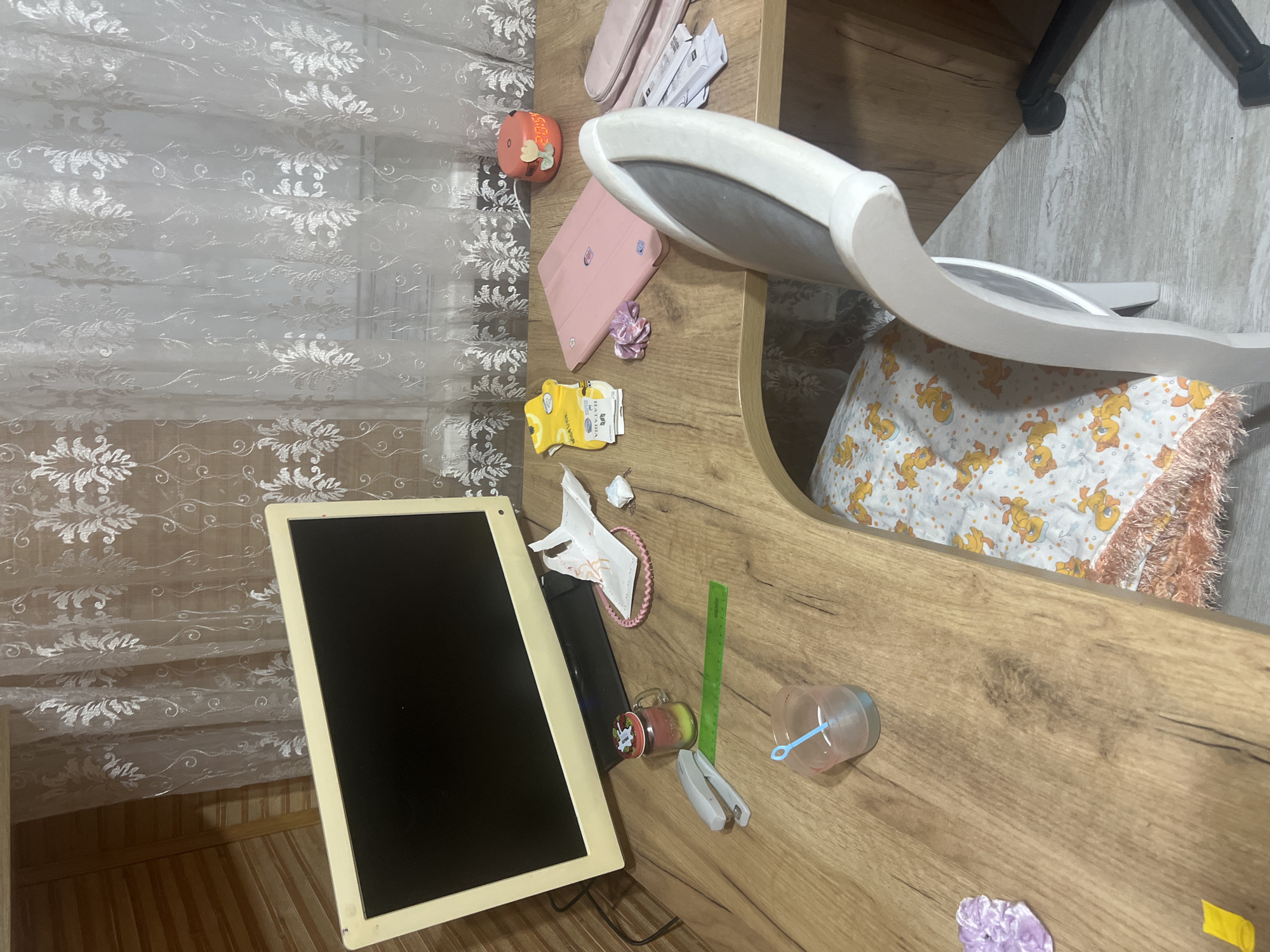 The image shows a wooden desk with various items, including a computer monitor, a stapler, a ruler, a cup with a blue stirrer, a jar with colored liquid, a pink hair scrunchie, a small piece of crumpled paper, a package of yellow socks, a pink tablet case, a small orange humidifier, and a pink makeup bag. Behind the desk are white lace curtains and a wooden wall. A white chair with a gray upholstered seat is positioned at the desk. The floor is light gray and wooden. Under the desk, part of a bed with a blanket featuring a cartoon duck pattern is visible. The scene appears to be in a domestic setting. The specific location within Surgut, Russia, cannot be determined from the provided image.