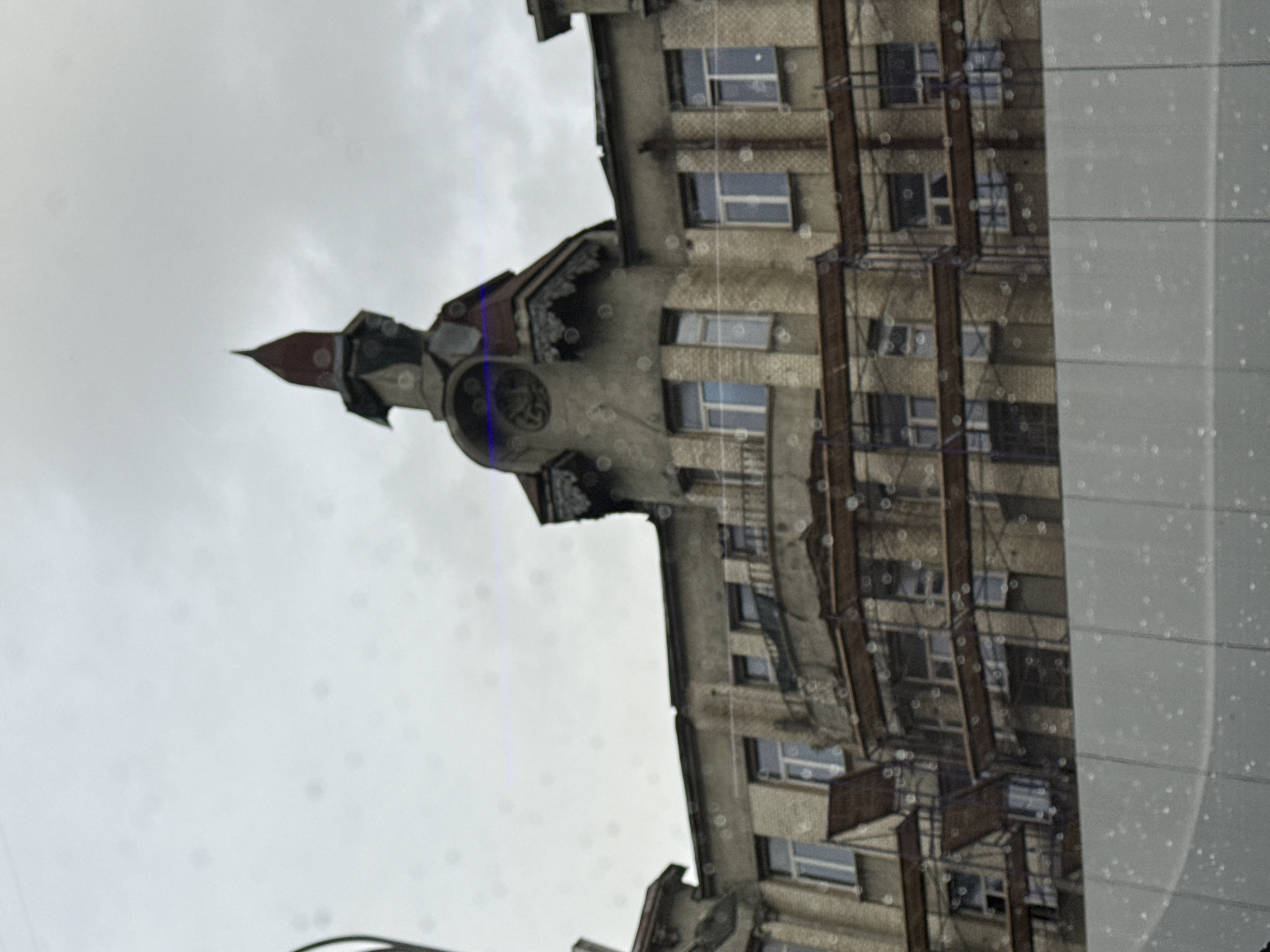 The image depicts the damaged Moscow House of Soviets, located in Moscow, Russia. The building is characterized by its multi-story structure and prominent tower element. Scaffolding is visible on the building. The weather appears overcast, and there are water droplets on the glass obscuring the view.