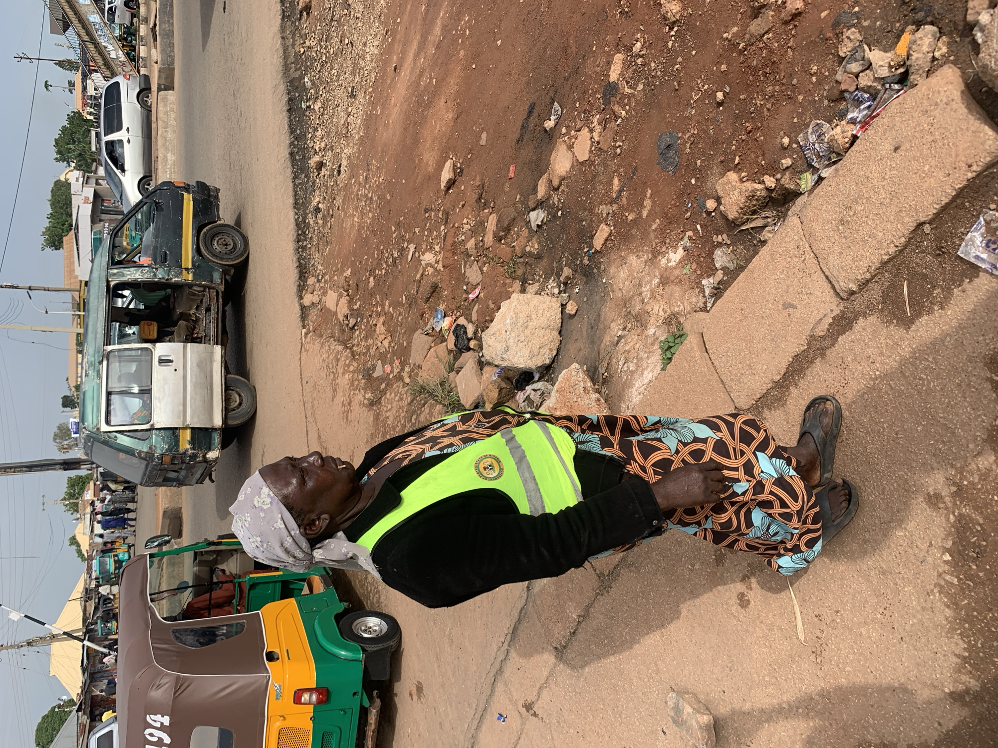 In Gura Top, Nigeria, a woman stands on a dirt and concrete roadside. She is wearing a black jacket over a patterned dress, a light blue and grey headscarf, and dark sandals. A bright neon green vest with reflective stripes is worn over her jacket. Her right hand is resting on her hip. Beside her, a green and yellow tricycle, known locally as an "okada" or "motorcycle taxi," is parked. The tricycle has the number "194" visible on its brown roof. In the background, a weathered green and white passenger van is stopped on the road, with its door open and people visible inside. Other vehicles and buildings can be seen further in the background, suggesting a busy urban or semi-urban environment. The ground is uneven, with a mix of packed dirt, loose stones, and scattered debris, including what appears to be plastic waste. A concrete curb runs along the edge of the road. The sky is overcast.