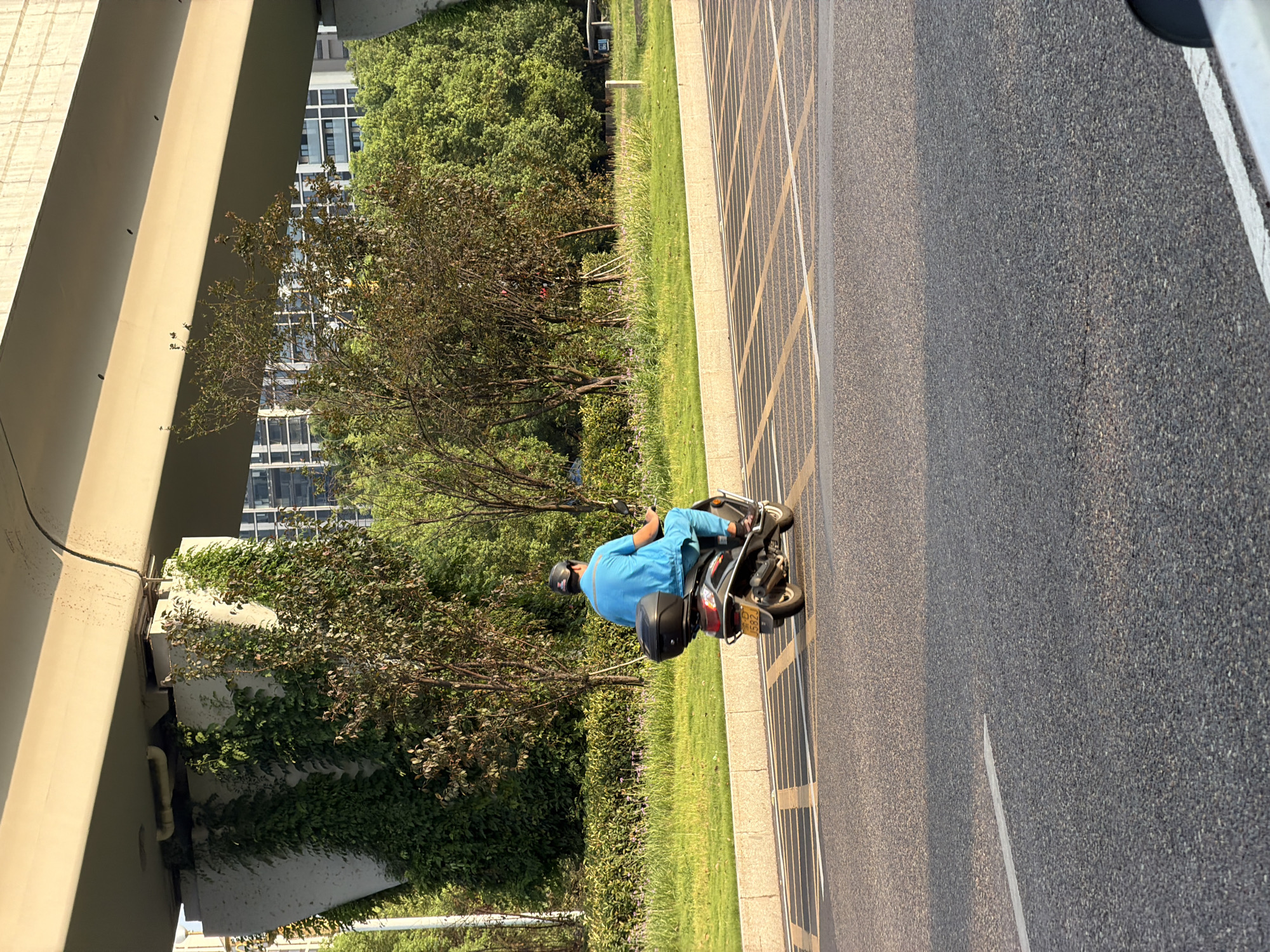 A person wearing a blue uniform and a black helmet is riding a scooter on a paved road in Jiangjin, China. The scooter has a black storage box on the back. The person is facing away from the camera and appears to be moving from left to right. In the background, there is a grassy area with trees and a large concrete overpass. Buildings are visible in the distance. The road has yellow and white markings. The sun is casting shadows, indicating it is likely late afternoon.