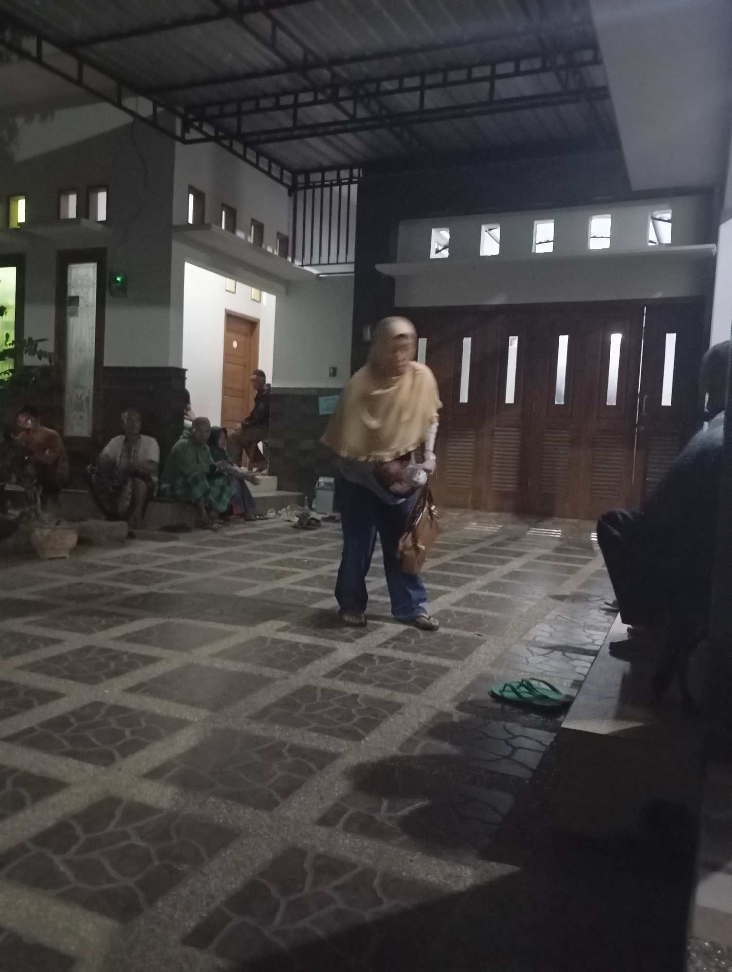 The image depicts an outdoor scene at night, likely in Banjarsari, Indonesia. A group of people are gathered in what appears to be the courtyard or patio area of a building. The architecture suggests a residential or community building, with a covered walkway or overhang supported by a metal frame. The ground is paved with large square tiles arranged in a diamond pattern, some of which are illuminated by ambient light, creating shadows.
In the foreground, a person wearing a light-colored headscarf and blue pants stands facing away from the camera, holding a brown bag. To their left, several individuals are seated on steps or the ground. One person in the middle ground wears a green headscarf and is seated amongst others. Further to the left, more people are seated, with one person in a white top. A plant in a pot is visible to the far left.
In the background, a building with several windows and doors is visible. One door appears to be made of wood with vertical slats and rectangular glass panels. Another door is a lighter color and has a handle. A man is seen seated on steps in the background, wearing dark clothing.
On the ground near the right side of the image, a pair of green flip-flops are placed. The lighting is dim, indicating nighttime, with artificial lights illuminating the scene from the building and possibly overhead. The overall impression is of a casual gathering or a moment of rest.