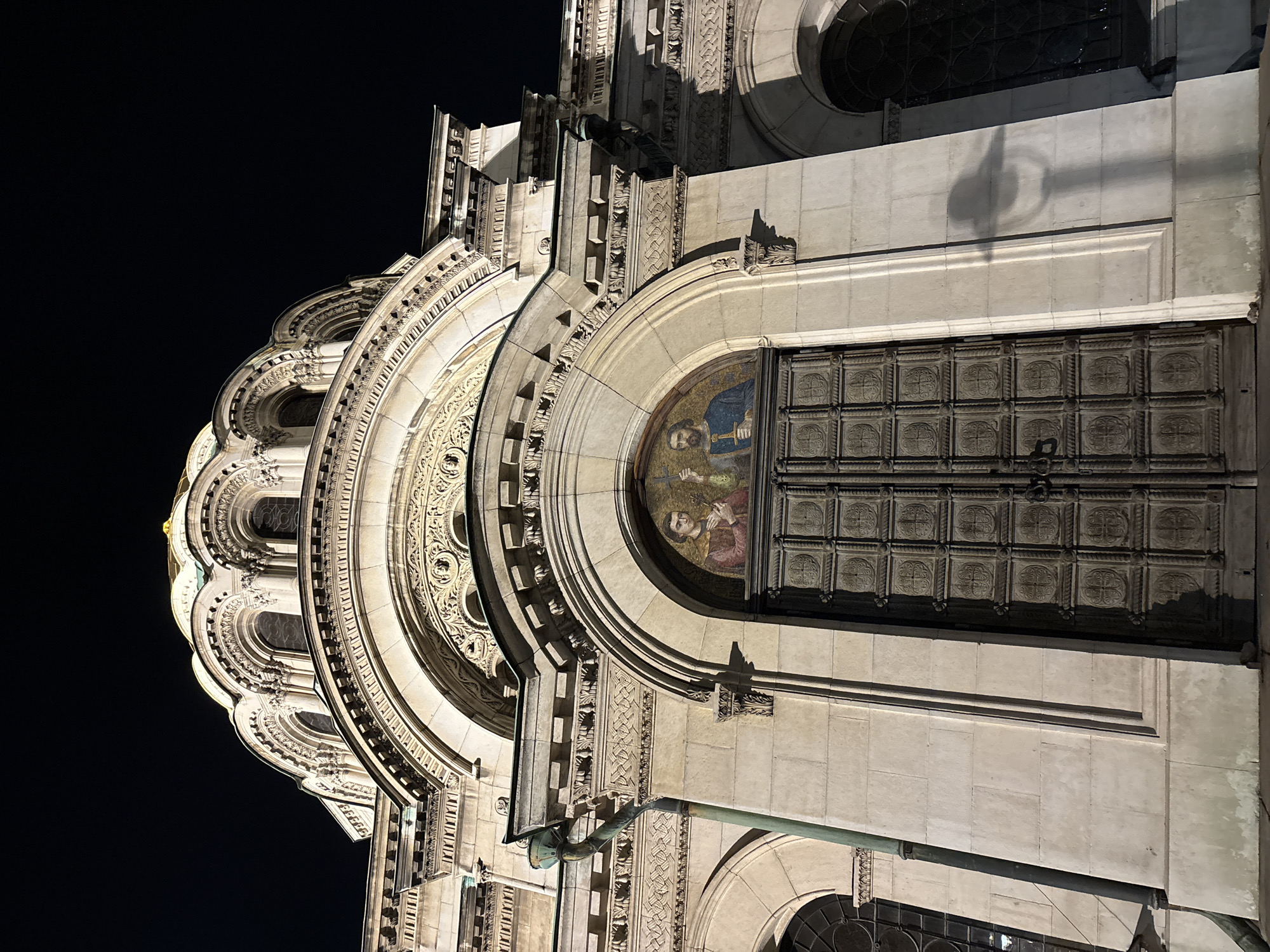 The image captures a dramatic, upward-angled view of the highly ornate facade of a grand, likely religious, building at night. The structure is brightly illuminated, standing out against a dark, clear sky.
In the foreground and center-right, a massive arched entrance dominates, featuring multiple layers of intricately carved arches adorned with decorative patterns. Above a large, dark, rectangular metal door, a colorful mosaic is embedded within the archway. The mosaic depicts three figures, likely religious, with two prominent bearded men; one in blue on the right holding an object resembling a cross or staff, and another below him in reddish-brown. The large door is composed of numerous square panels, each embossed with a decorative, possibly quatrefoil, design.
To the left, the building's intricate architecture continues, showcasing multiple tiers of arched windows, columns, and elaborately carved stone details. These elements recede into the distance, culminating in a glimpse of a curved, gold-colored dome or roof section on the far left. A metal downspout or pipe is visible on the lower right portion of the facade. The artificial lighting creates strong contrasts and shadows, highlighting the architectural depth and rich textures. There is no visible text in the image. The scene suggests a significant landmark, likely the Alexander Nevsky Cathedral, known for its Byzantine Revival architecture and mosaic details, illuminated in the night in Sofia, Bulgaria.