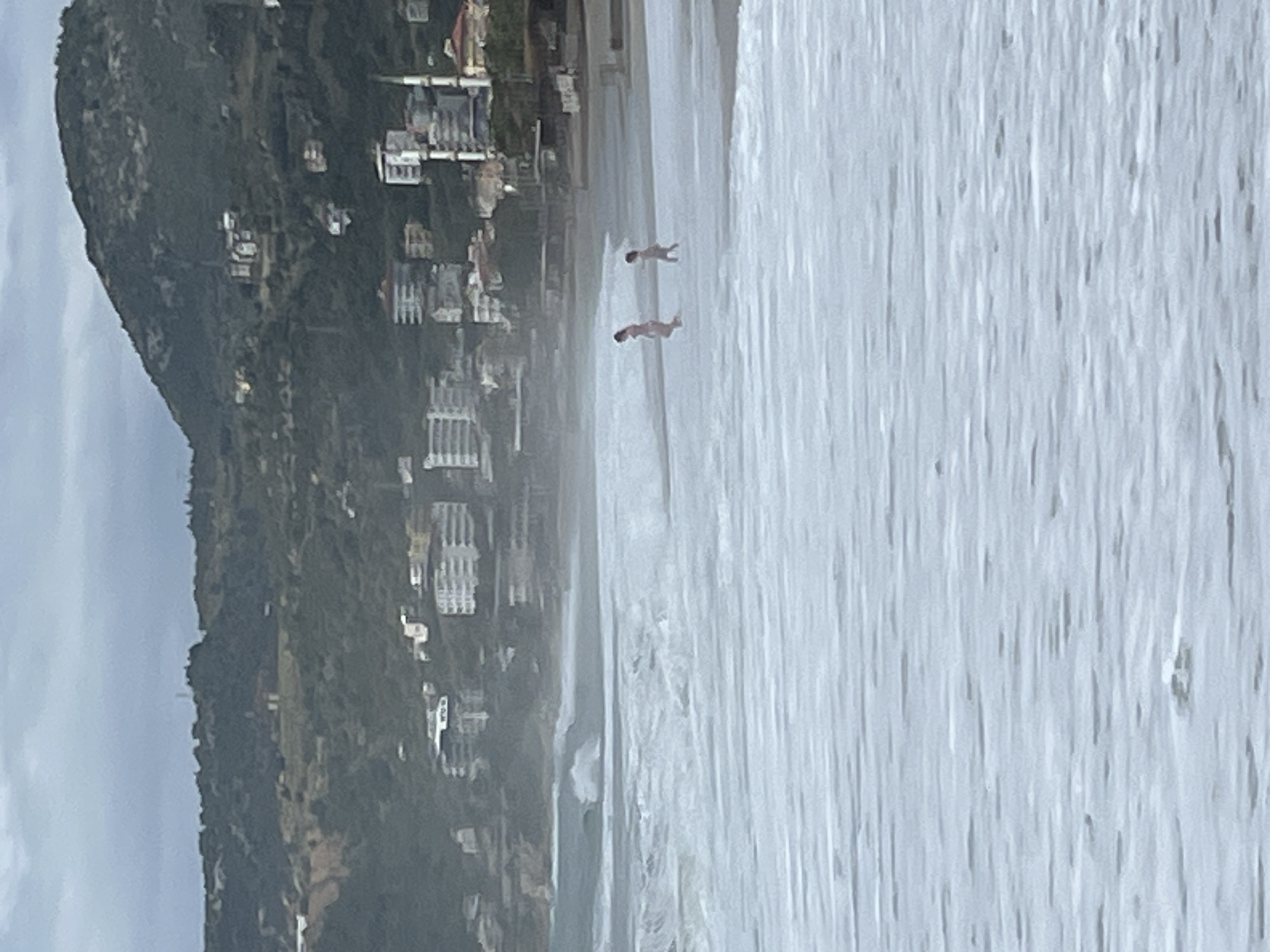 The image shows a coastal scene in Alanya, Turkey. A beach with white water and waves is in the foreground. Two people, possibly children, are wading in the water. Along the coast, there are buildings and a hillside covered in vegetation. The weather appears overcast.