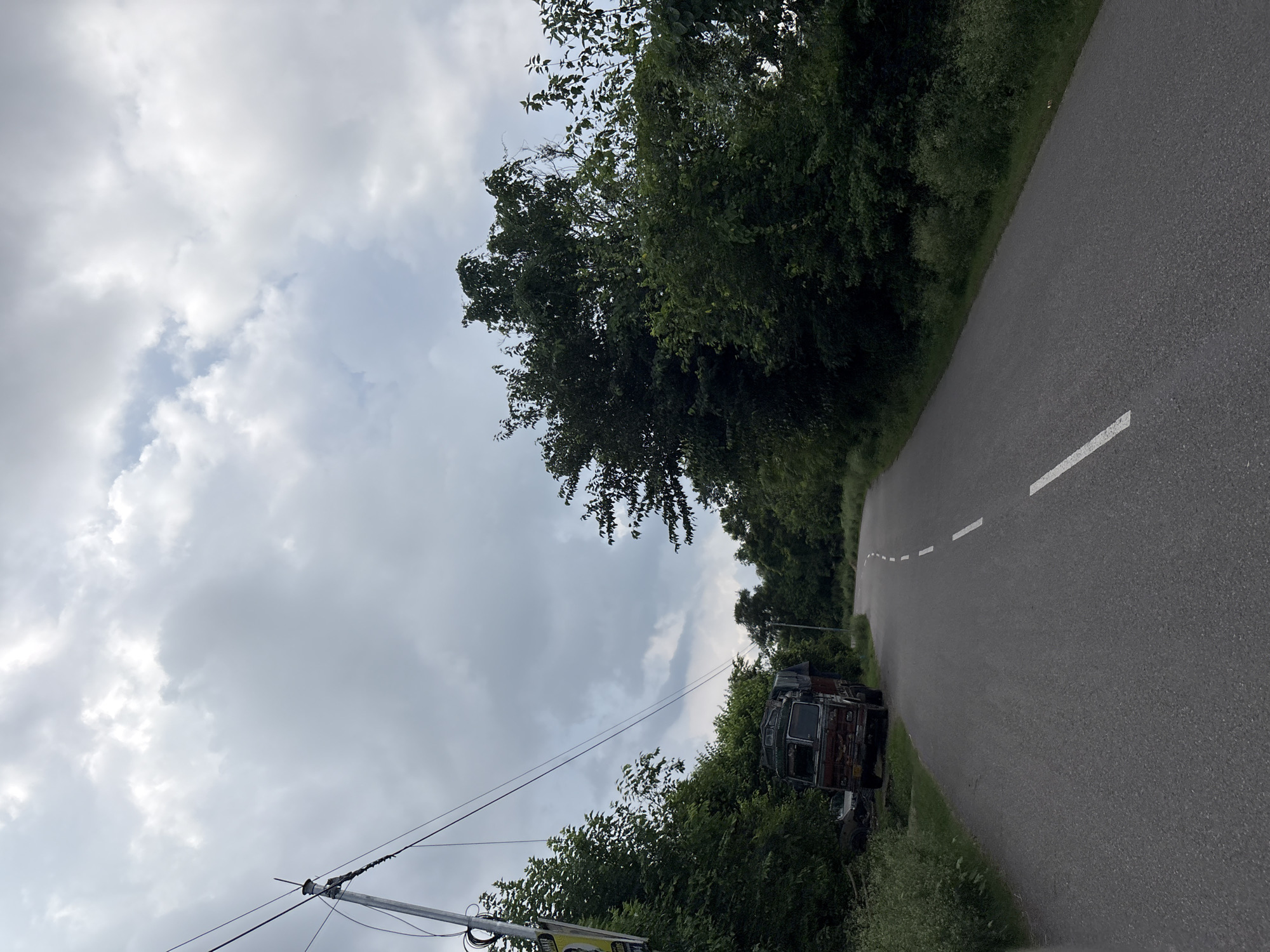 The image shows a slightly overcast sky over a rural road in Baldwara, India. A paved road curves to the right, marked with a dashed white line and solid white lines. On the left side of the road, a rusty, old truck is parked near some overgrown bushes and trees. There is also a utility pole with wires on the left side of the road, and a yellow sign attached to it. The right side of the road is lined with dense green trees and foliage. The overall scene suggests a quiet, somewhat aged rural environment.