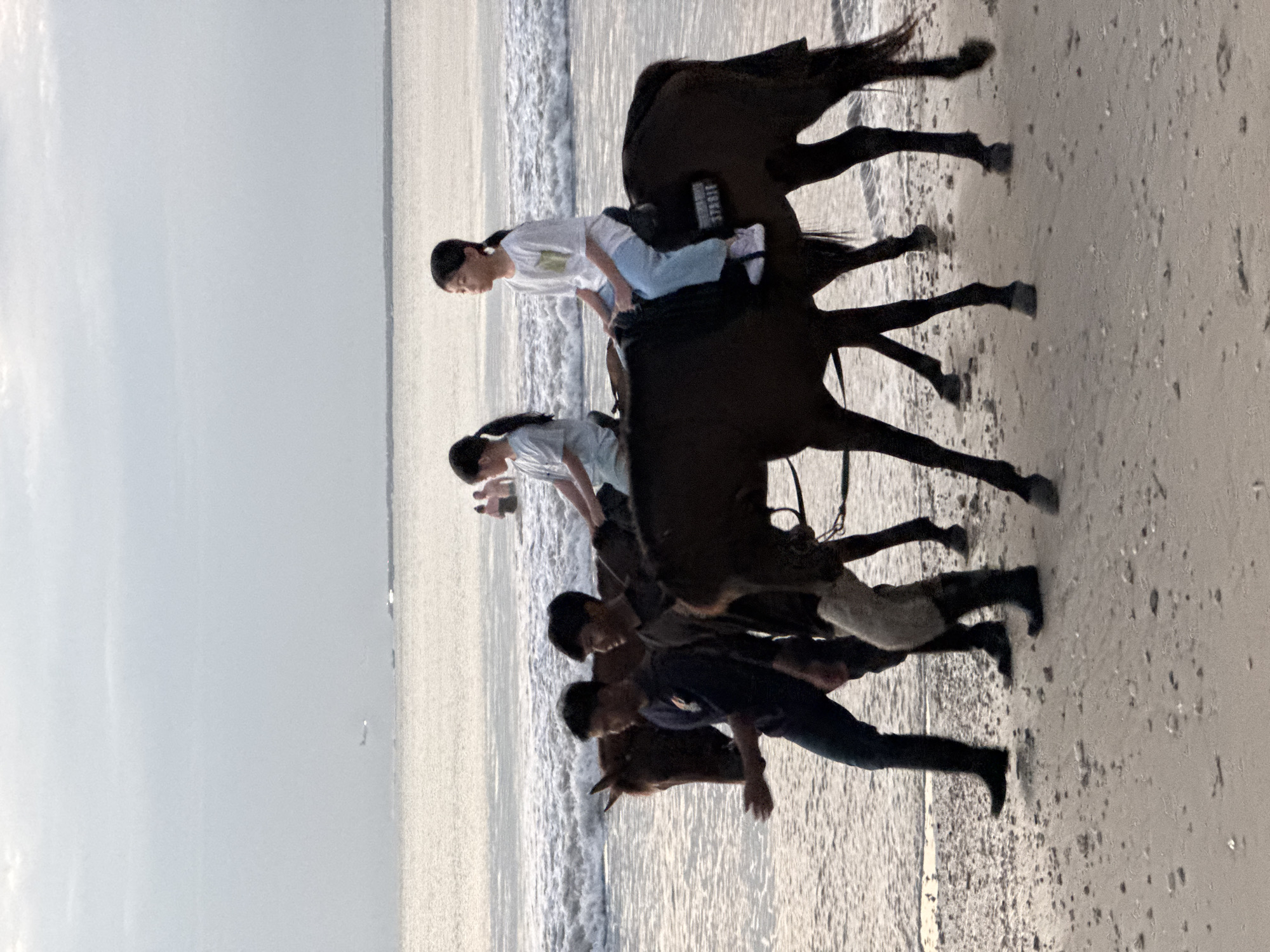 Two young girls, each on horseback, are being led along a sandy beach by two men. The girls are wearing light-colored t-shirts and pants, and the horses are dark brown. The men are dressed in dark shirts and pants, with one of them wearing boots. The horses are walking in the shallow water at the edge of the ocean, with waves gently lapping the shore. In the background, the ocean stretches out to the horizon, with a faint airplane visible in the sky. The overall scene appears to be a peaceful and leisurely activity on a beach in Kuta Selatan, Indonesia.