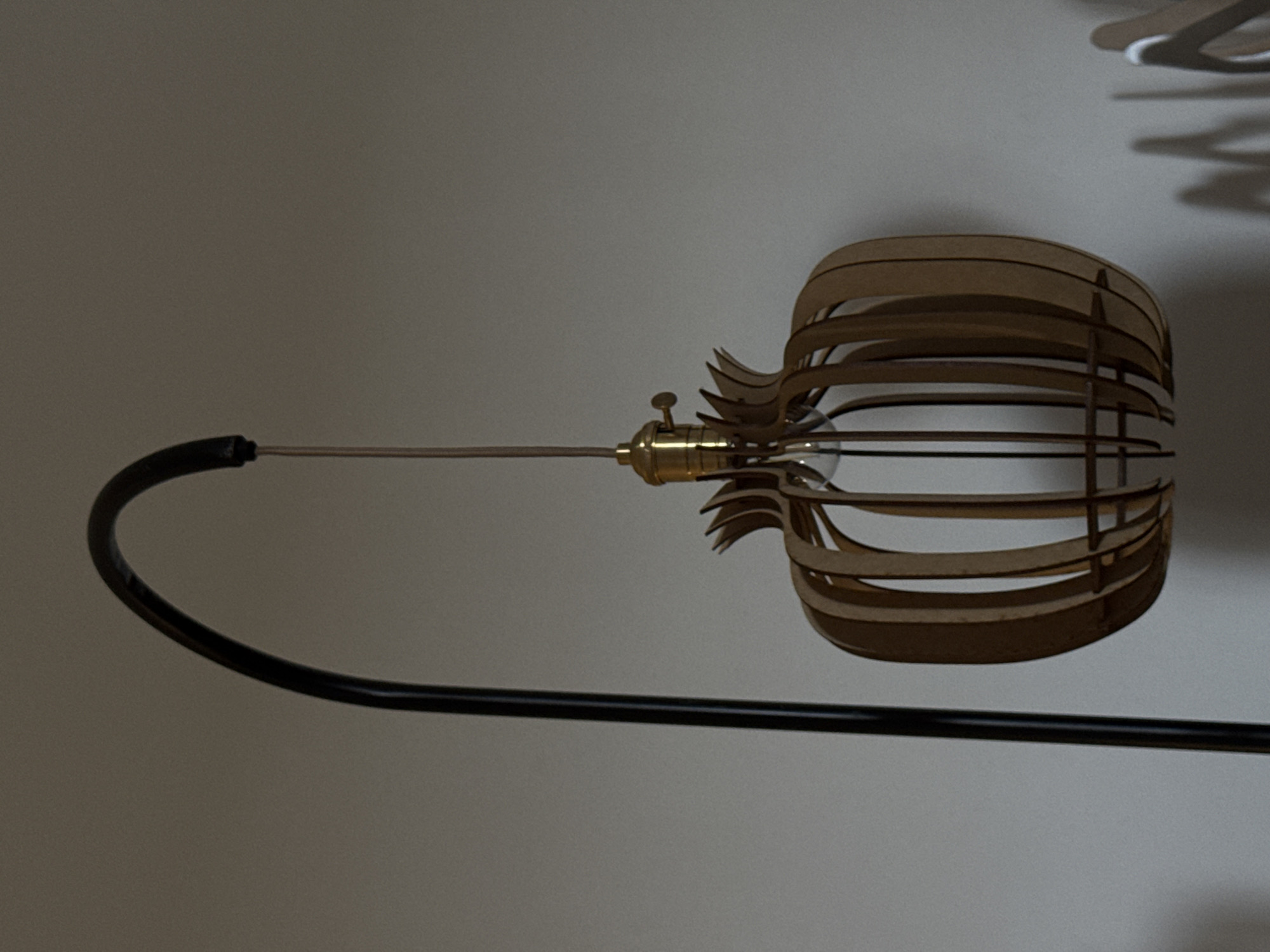 The image captures a close-up of a modern lamp against a plain, light-colored wall in San Andrés Cholula, Mexico. The central subject is a unique light fixture suspended from a curved, black metal arm, which appears to be part of a floor lamp. The fixture itself consists of a brass-toned metal socket holding an Edison-style light bulb with visible filaments.
Encasing the bulb is an intricate lamp shade made from numerous thin, light-colored wooden or composite slats. These slats are arranged in a complex, curvilinear, and layered pattern, forming a decorative, cage-like structure that adds an organic and artistic element to the lamp's design. The natural wood grain of the slats is discernible, enhancing the lamp's aesthetic.
The background is a uniform, muted light gray or off-white wall, providing a clean and understated setting that draws attention to the lamp. Soft, diffused shadows, likely cast by parts of the lamp or other objects outside the frame, are visible on the wall in the upper right portion of the image. No people or specific activities are depicted. The scene is static, highlighting the lamp as a decorative and functional object within an indoor space. There is no visible text in the image.