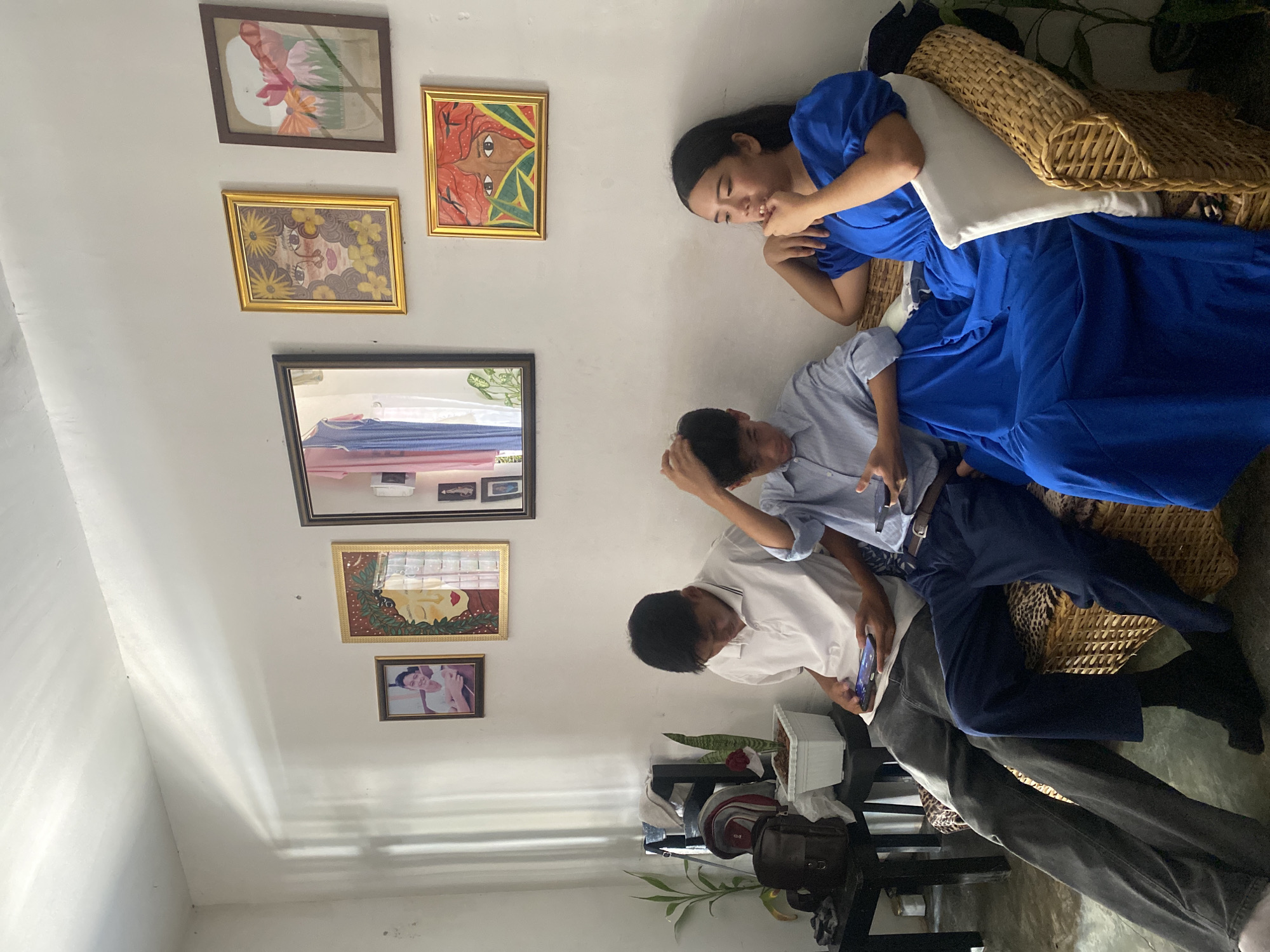 This image, captured in Davao City, Philippines, depicts three young individuals seated on a wicker sofa with leopard-print cushioning, positioned against a white wall adorned with framed art. On the far left, a male in a white polo shirt and dark jeans is focused on a smartphone he holds in both hands. Adjacent to him, a second male, wearing a light blue collared shirt and dark blue trousers, also holds a smartphone, his right hand lightly touching his hair. To the right, a female dressed in a bright blue dress sits with her left hand near her mouth, gazing slightly to the left with her lips parted. The white wall features an arrangement of six framed items: a small framed photo, two framed artworks depicting stylized faces, a large rectangular mirror reflecting interior details, and two framed floral/abstract artworks. Streaks of natural light are visible on the upper left portion of the wall. To the left of the sofa, a dark table holds a small backpack, a dark shoulder bag, and two potted plants, one resembling bamboo and the other a snake plant in a white pot.