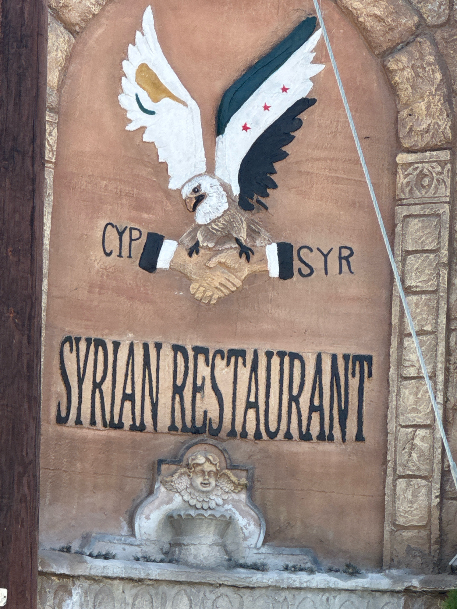 A stucco relief sign is mounted on a weathered, tan-colored building facade, framed by rough-hewn stone archways and decorative stone pilasters. At the top of the sign, a stylized eagle with outstretched wings, painted white, green, and black with red stars on its wings, hovers above two clasped hands. The wings appear to represent the Syrian flag. Text in black capital letters above the clasped hands reads "CYP" to the left and "SYR" to the right, suggesting Cyprus and Syria. Below the eagle and hands, larger black capital letters spell out "SYRIAN RESTAURANT". At the bottom of the sign, a decorative relief of a cherubic face with feathered wings emerges from a stone structure. The sign is mounted outdoors, with a thin wire extending diagonally from the upper right to the lower right. A portion of a dark wooden pole is visible on the far left edge of the image. The location is identified as the Municipality of Germasogeia, Cyprus.
