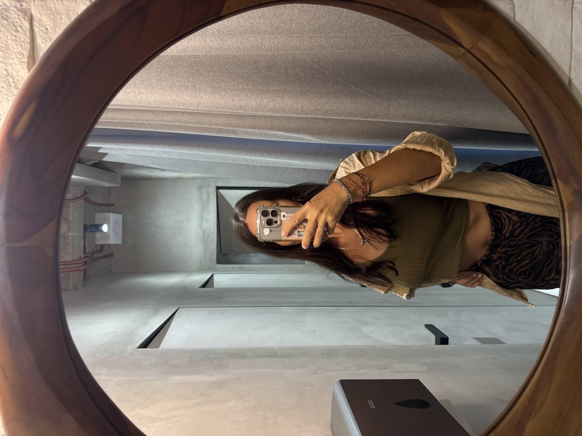 The image displays a mirror selfie, with the entire composition rotated 90 degrees counter-clockwise. A person is reflected in a large, circular mirror featuring a substantial, polished wooden frame. The individual appears oriented on their side within the reflection, holding a smartphone with their left hand to capture the image; their face is largely obscured by the phone.
The person has long, dark hair and is dressed in a light, neutral-toned, long-sleeved shirt or jacket worn open over a ribbed green crop top. Their lower half is clad in black and brown zebra-print pants. Multiple delicate bracelets adorn their right wrist, and rings are visible on the fingers of their left hand. The smartphone features a clear case embellished with small floral patterns around its triple camera setup.
The reflected background reveals a modern interior space, possibly a public restroom or contemporary establishment. The walls and ceiling are light gray, showing a textured, concrete-like finish. Along one wall, exposed copper piping runs beneath a white rectangular light fixture that projects light. The floor, also light gray, is characterized by sharp, dark, linear recesses that suggest architectural design elements or vents. A dark gray or black dispenser, likely for soap or sanitizer, is partially visible in the lower part of the foreground. There is no clearly discernible text. The scene is brightly lit by artificial indoor lighting, consistent with a contemporary venue in Kuta Selatan, Indonesia.