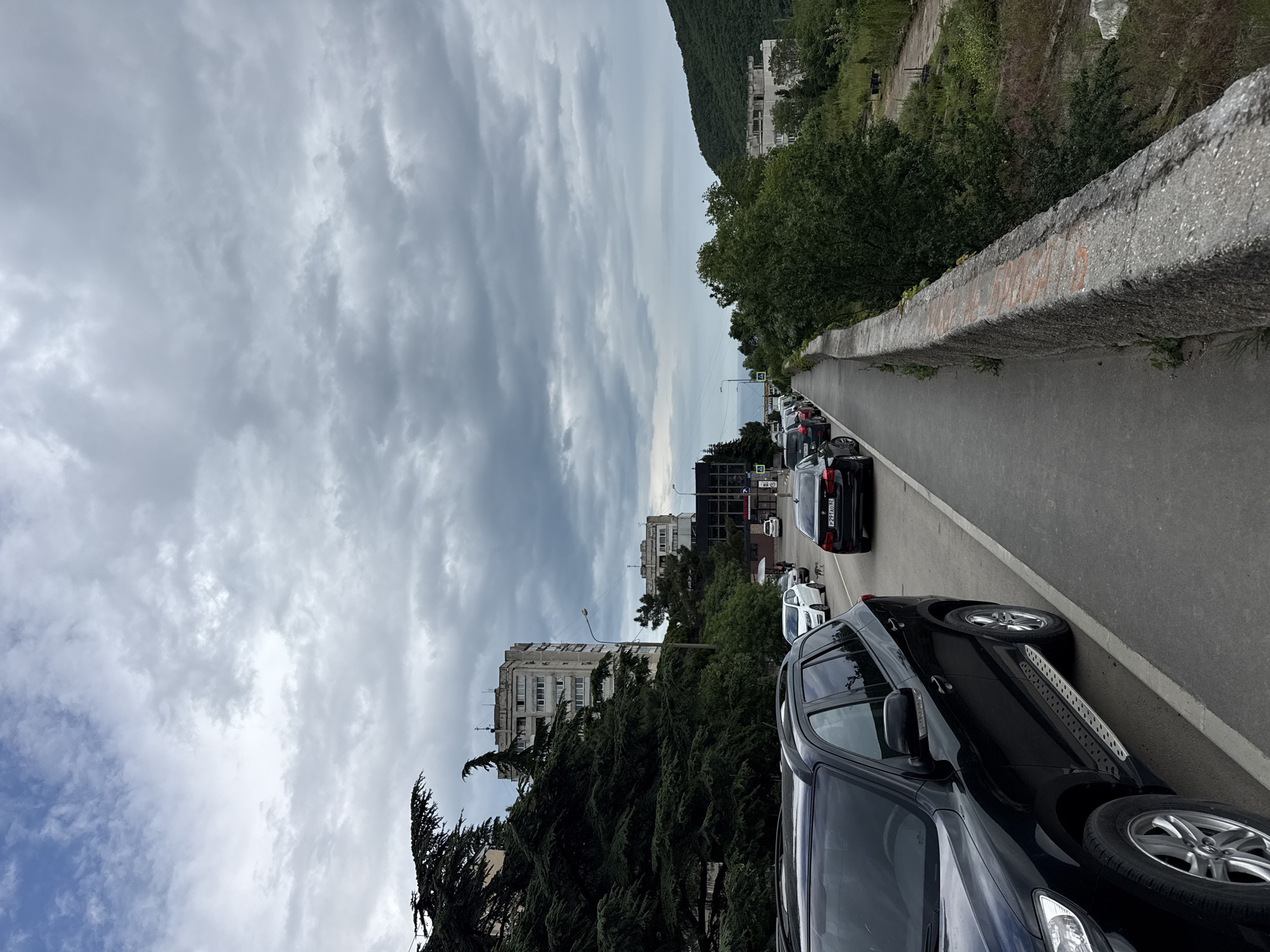 A street scene, possibly a bridge, in an urban area. Numerous cars are parked along the right side of the street, and a concrete wall runs along the edge of the road. The sky is overcast. Several buildings are visible in the background, as is a tree-covered hillside.