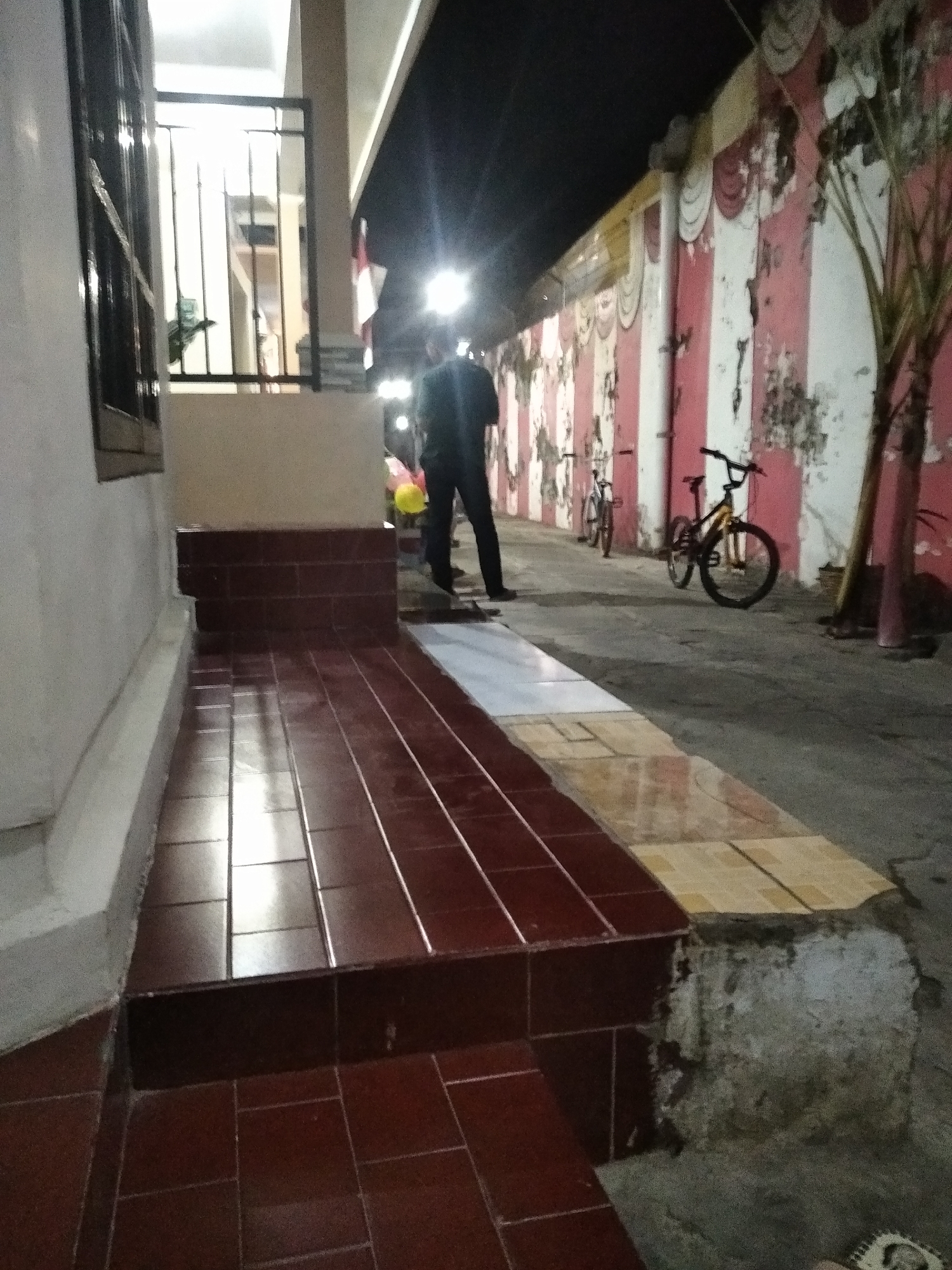 The image depicts an outdoor scene at night, likely in Rambipuji, Indonesia, with a man standing near a building entrance. The man, dressed in dark clothing, is partially visible on the right side of the frame, facing away from the camera. In front of him, a yellow balloon is held by an unseen individual.
The foreground features a tiled walkway with dark red and lighter beige tiles. A raised structure, possibly a step or platform, is clad in dark red tiles, with a white concrete base. To the left, a portion of a building wall with a dark window frame is visible.
In the background, a wall is decorated with alternating pink and white vertical stripes, exhibiting signs of wear and peeling paint. Two bicycles are parked along this wall. The environment is lit by artificial lights, creating reflections on the wet-looking tiled surfaces. The overall impression is that of a residential or public space at dusk or night, possibly during an event or gathering.
