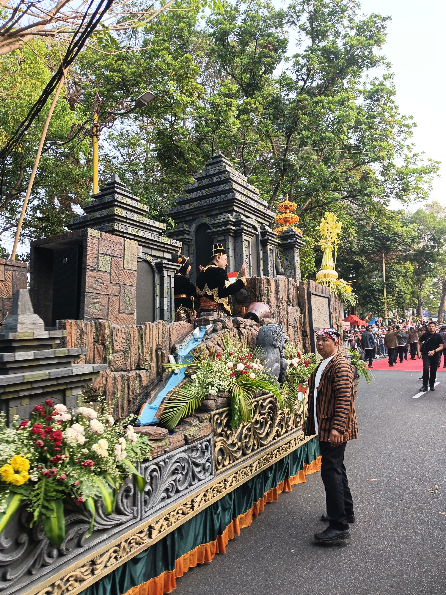 The image captures a scene from a parade in Gayamprit, Indonesia. In the foreground, a man wearing a striped jacket, white t-shirt, and black pants stands on a street, facing right. He is looking slightly upwards and to the right, with a neutral expression. To his left is a decorated float.

The float is elaborate, designed to resemble a stone structure with steps and a waterfall feature. It is adorned with lush green plants and a variety of flowers in red, pink, and yellow hues. Behind the waterfall, two figures dressed in traditional attire, possibly representing a historical or cultural performance, are seated. One figure is wearing a black outfit with gold trim and a traditional headdress, while the other is similarly dressed in black and gold. Further back on the float, a golden ornamental structure stands prominently.

The background reveals a street lined with trees, their green leaves creating a canopy. A crowd of people is visible further down the street, suggesting a public event or festival. Some individuals are dressed in traditional Indonesian clothing, while others wear casual attire. Streetlights are mounted on poles along the street. The overall atmosphere appears festive and ceremonial.
