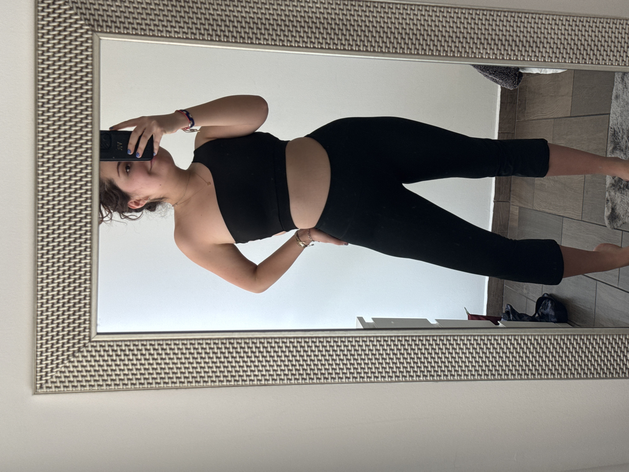 A person in a black top and black leggings is standing in front of a mirror, taking a photo of themselves with a cell phone. The mirror has a silver-colored frame with a textured pattern. The background visible in the mirror includes a white wall, a tiled floor, and some clothing items. The location appears to be a room inside a building. There is no indication of the city of Mexico City, Mexico in this image.
