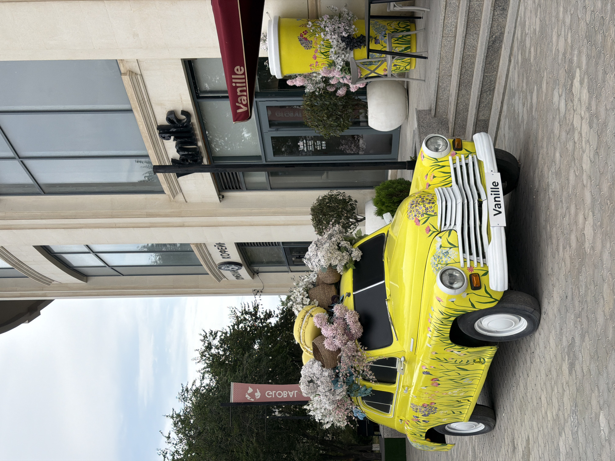 A vintage yellow car, adorned with floral decals and a large macaron on its roof, is parked on a paved walkway in front of a light-colored building. The car is decorated with an abundance of white and pink flowers. The building features large windows and an awning with the word "Vanille" in white lettering. To the right of the car, a giant yellow coffee cup with floral designs and a small table with chairs are visible. The background includes trees and a cloudy sky. The scene is located outside a "Vanille" cafe on the Caspian Sea embankment in Aktau, Kazakhstan.