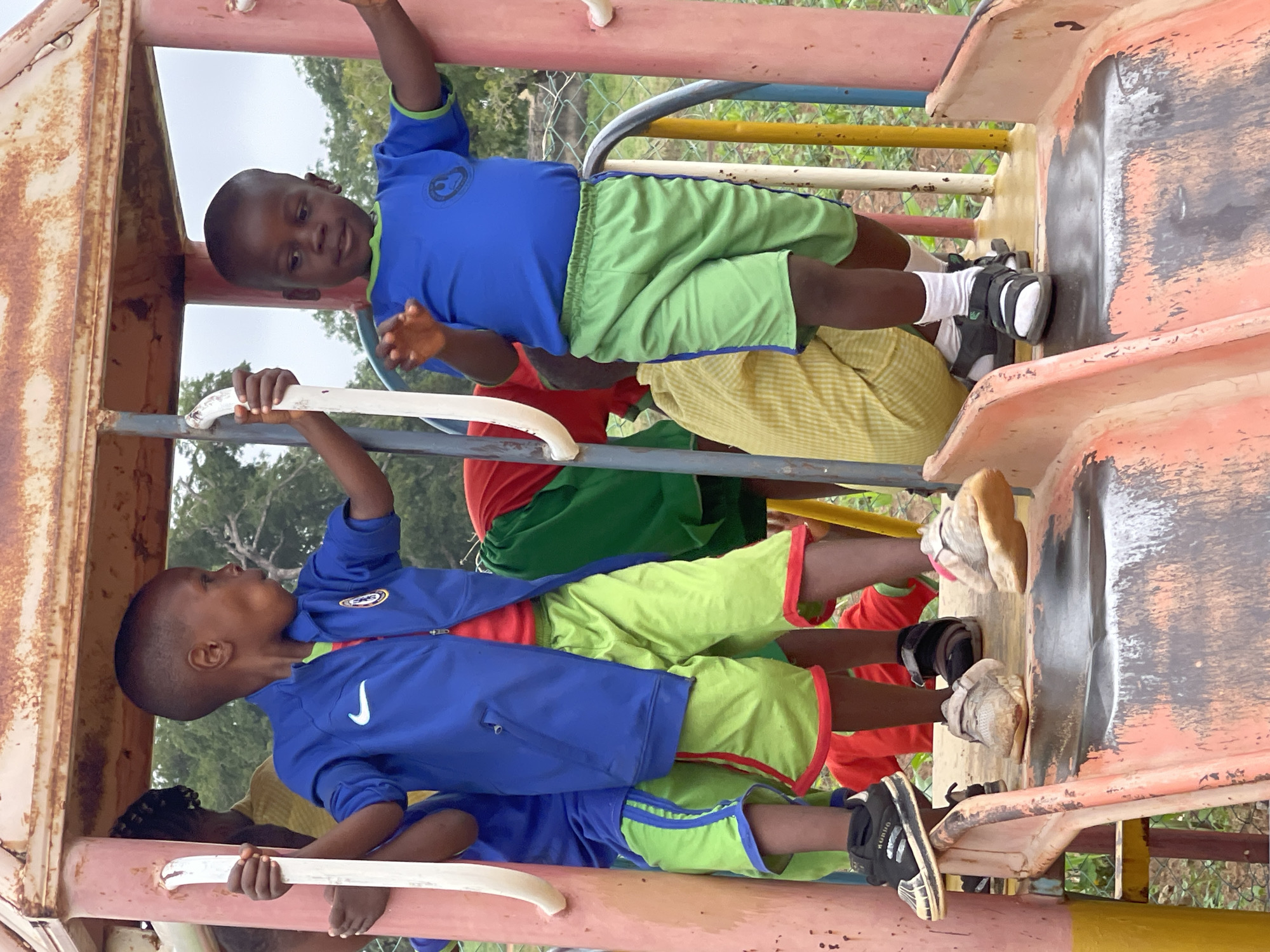 This image captures a vibrant scene of children playing on a well-worn playground structure in Akwanga, Nigeria, during what appears to be daytime under an overcast sky.
Multiple young children, likely boys, are actively engaged with the rusty, multi-colored playground equipment. In the upper portion of the frame (when oriented correctly), a boy wearing a blue long-sleeved shirt with a circular emblem and green shorts is positioned at the top of a slide or climbing frame. Notably, he wears a black and white medical brace or boot on his right foot, along with white socks and shoes. He looks towards the viewer with a focused expression.
Below him, another boy in a blue zippered jacket featuring a white Nike swoosh logo and green shorts holds onto a white horizontal bar, looking upwards. Further down, parts of other children are visible; one is in a red shirt and green shorts, and another with braided hair is near the bottom of the frame, also holding a white bar. The children’s clothing is bright, contrasting with the faded and peeling paint of the playground.
The playground structure itself shows significant signs of age and use, with rust and chipped paint in shades of orange, red, and brown on the slide and supports. The climbing bars are painted in faded white, blue, yellow, and red. In the background, green trees and foliage are visible, suggesting a natural outdoor setting, possibly with a fence, under a bright, diffused sky. The children appear to be engrossed in their play, showcasing resilience and joy in their activity. Partial text "KUAO" is visible on one child's shoe.