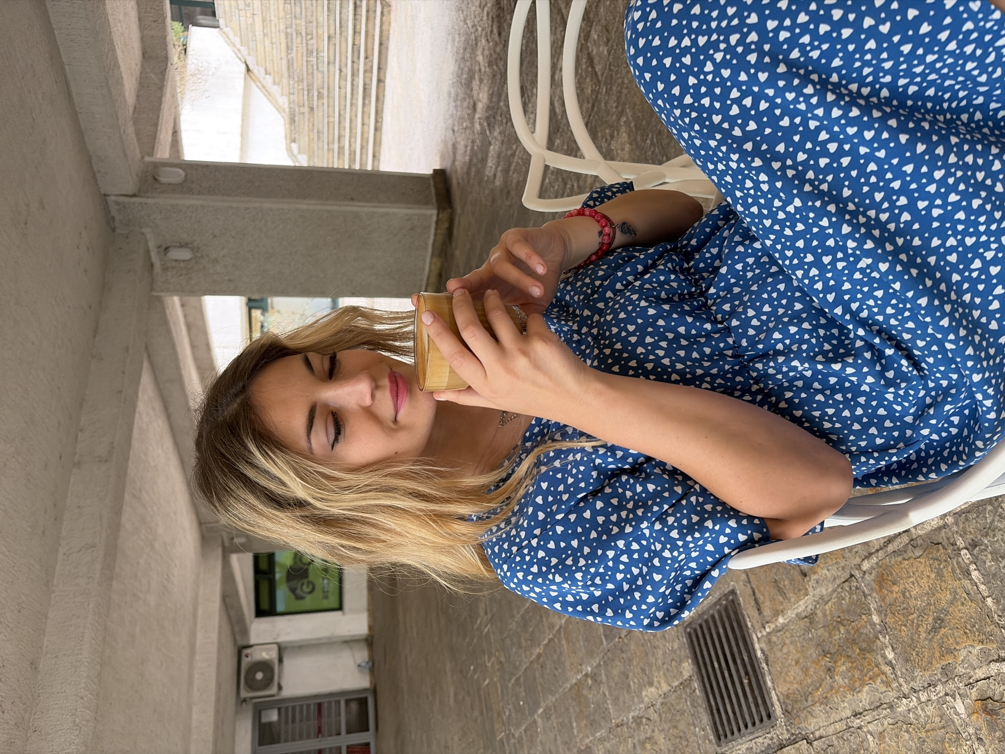 The image, taken in Budva, Montenegro, shows a young woman seated outdoors during the daytime, enjoying a beverage. She has long, wavy blonde hair and wears a blue dress with a white heart pattern. With a serene expression, eyes partially closed and a slight smile, she holds a clear glass cup containing a layered drink, likely a coffee, with both hands. A red beaded bracelet and a small dark tattoo are visible on her left wrist.
She is seated on a white chair, part of which is visible in the foreground. The ground is paved with light-colored, textured stone or flagstones, with a metal grate embedded in it. In the background, there are robust light-colored concrete beams or architectural structures, possibly part of a building's foundation or an overhead passage. Further back and to the right, concrete stairs ascend. On the left, a white wall features an external air conditioning unit and a window with a green frame, displaying some faint, illegible text. The scene conveys a relaxed atmosphere in a bright, sunny outdoor setting.