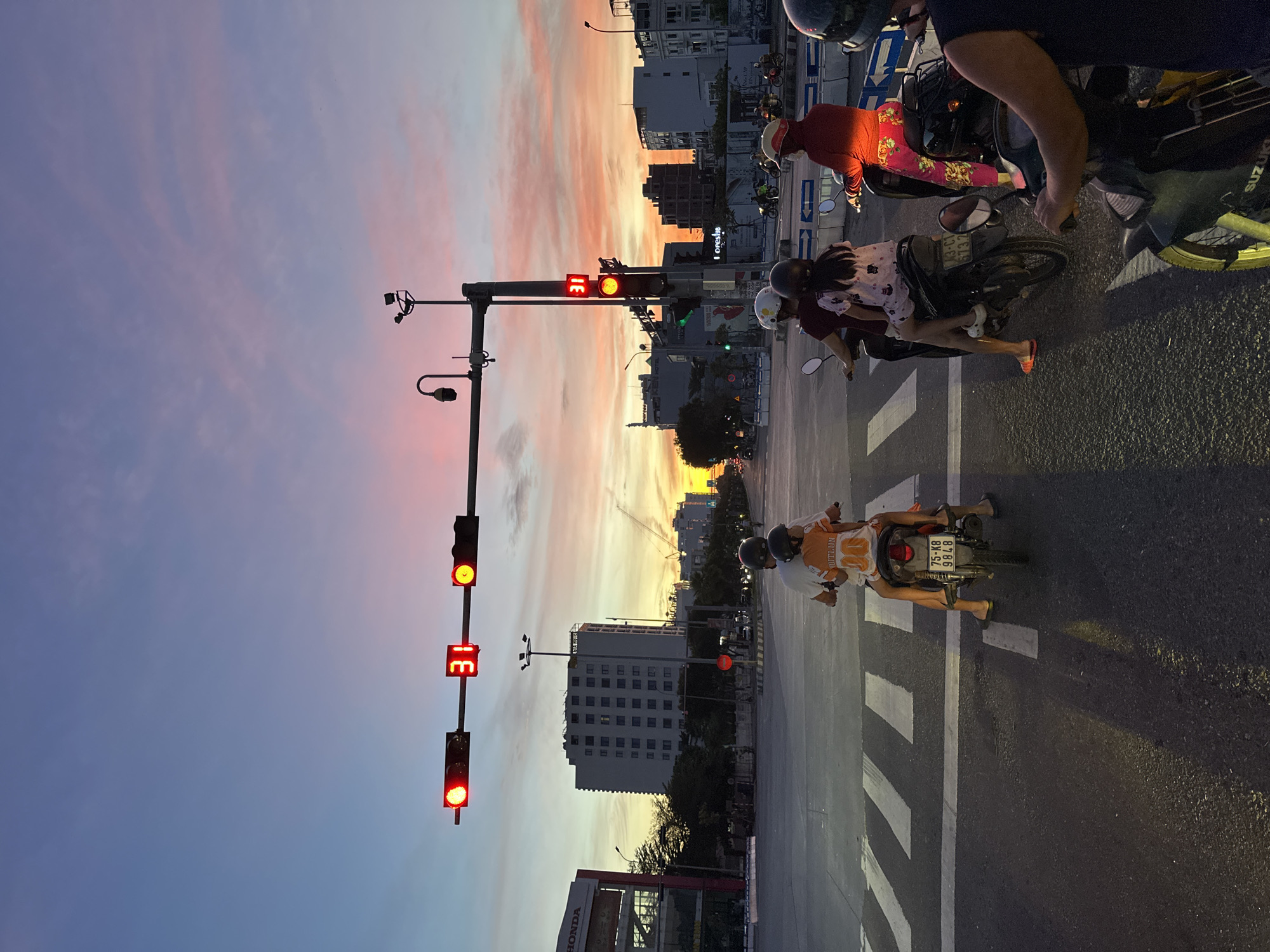 This is a full, eye-level shot of a street intersection in Da Nang, Vietnam, at dusk. The sky is a gradient of deep blue at the top, transitioning to soft pinks and oranges, indicating the setting sun. A traffic light with a red and yellow light illuminated is prominently displayed in the center of the frame. The digital display on the traffic light shows "31". Several motorcycles are stopped at the intersection, waiting for the light to change. On the leftmost motorcycle, a person wearing a white shirt and a child in an orange shirt with the number "10" are seated. A second motorcycle to their right has a person wearing a burgundy shirt and a child in a pink and white patterned outfit. A third motorcycle further to the right shows a person wearing a red hooded jacket and pink patterned pants. In the foreground, a person on a motorcycle wears a dark blue shirt and a black helmet. Buildings of varying heights line the street in the background, some appearing to be residential or commercial. A Honda dealership sign is visible on the left. The road surface is asphalt, with white pedestrian crossing lines painted on it. The overall scene depicts a typical urban moment in Da Nang during sunset, with traffic and urban infrastructure under a colorful sky.