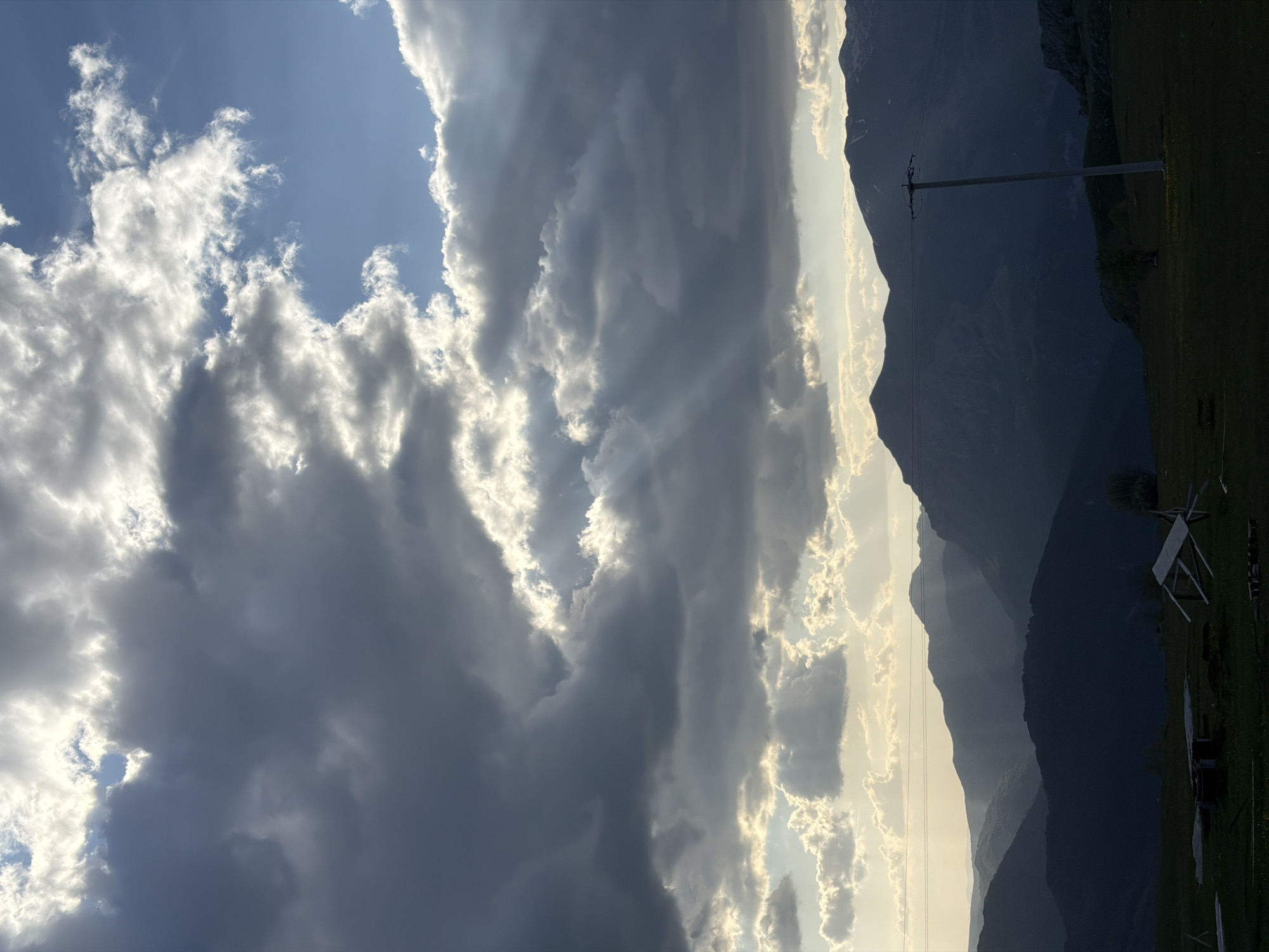 The image shows a landscape of mountains and clouds in Pišče, Montenegro. The left side of the image is dominated by large, heavy clouds, with some patches of blue sky visible. To the right, a silhouette of mountains is seen with sunlight filtering through, indicating the position of the sun. An electrical post is visible on the right side of the mountains. A grassy area is visible at the bottom right, suggesting a field or meadow.