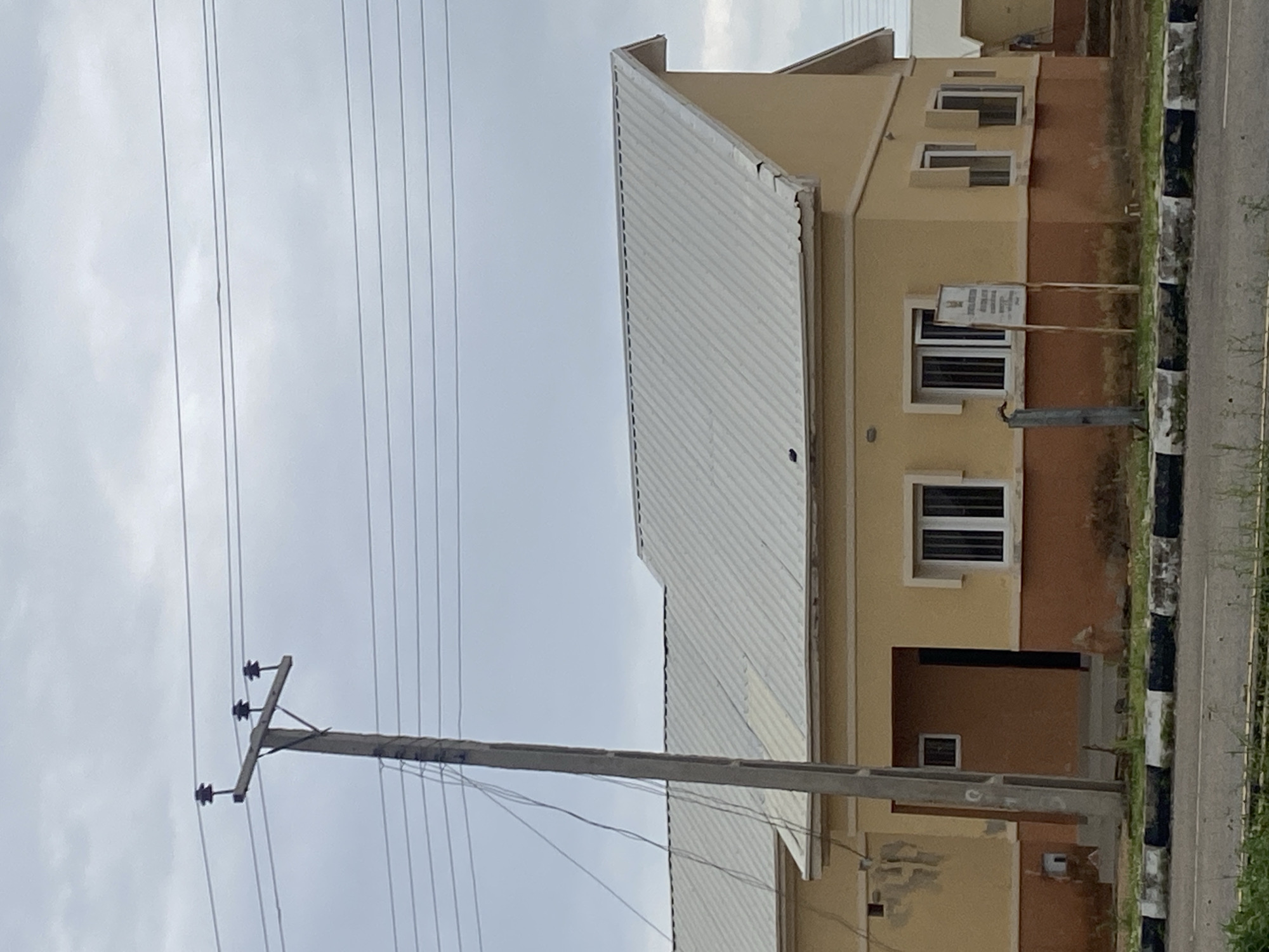This image captures a view of a residential building in Bulamare, Nigeria, under an overcast sky. A concrete utility pole stands prominently in the foreground to the left, supporting multiple power lines that stretch across the frame. The building features a light yellow facade with a darker orange-brown wainscoting. It has a light-colored, corrugated metal roof with a distinctive angled peak on the right side. Several windows with white frames are visible, some with visible bars. A signpost with text is located near the right corner of the building. The building is situated behind a curb with alternating black and white markings, beyond which a paved road is visible. Green vegetation, including grass and some weeds, is present along the side of the road and in front of the building. The overall scene depicts a typical residential setting in a Nigerian urban or suburban environment.