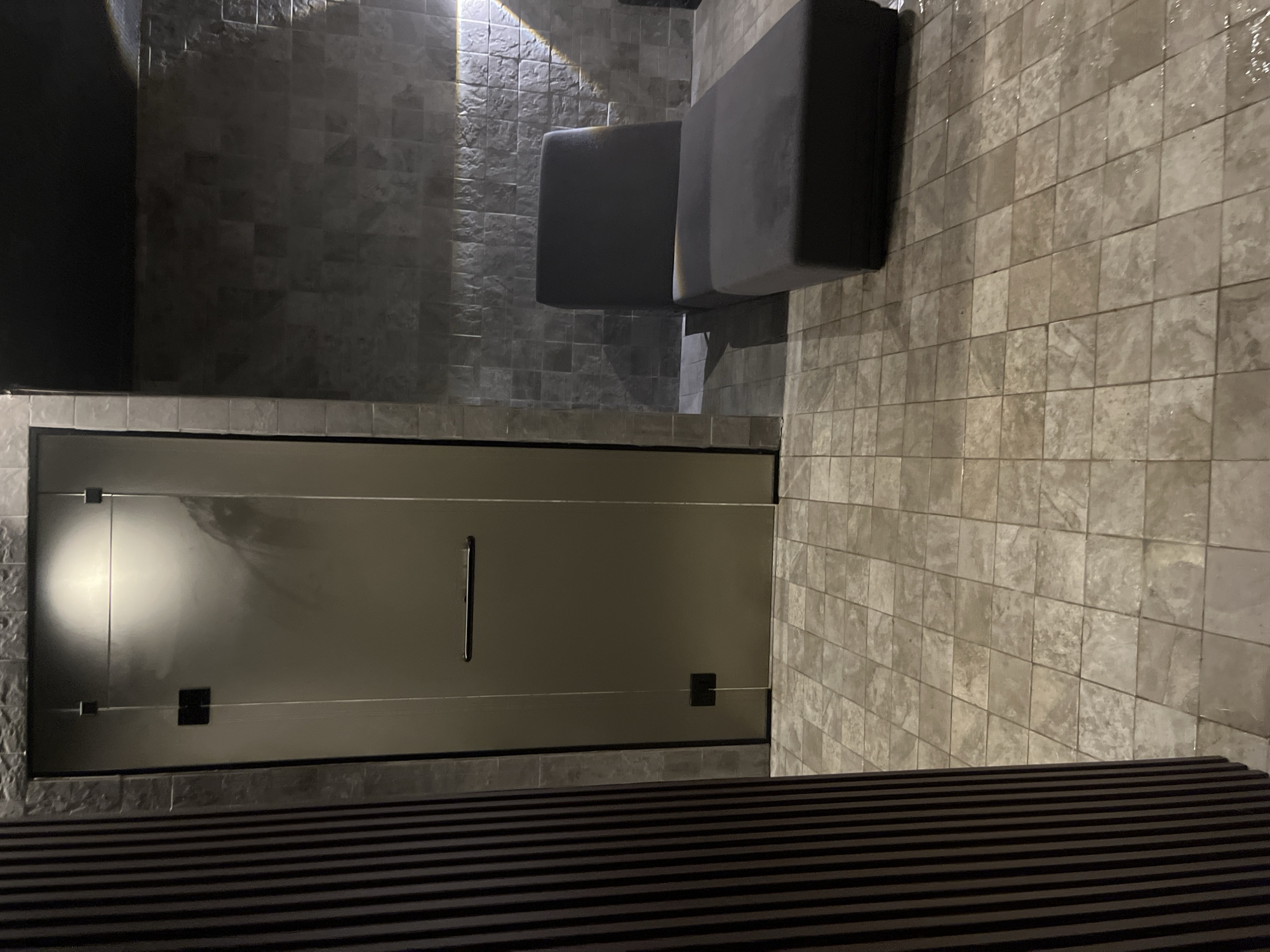 The image shows a room with gray tiled walls and floor. There is a glass door with a handle in the middle of the frame. Above the door are two gray ottoman-like seats. At the bottom of the image are dark horizontal stripes. There are no people visible. The location is unknown.