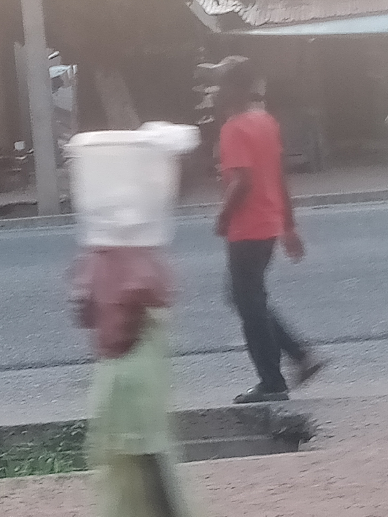 In Jalingo, Nigeria, two individuals are visible on a paved surface adjacent to a road. In the foreground, a person in a light green garment walks from left to right, carrying a large, white, translucent container on their head. Behind and to the right of this person, another individual, wearing a red t-shirt and dark trousers, walks in the same direction. The background shows a blurred street scene with structures and foliage. The lighting suggests it is daytime.