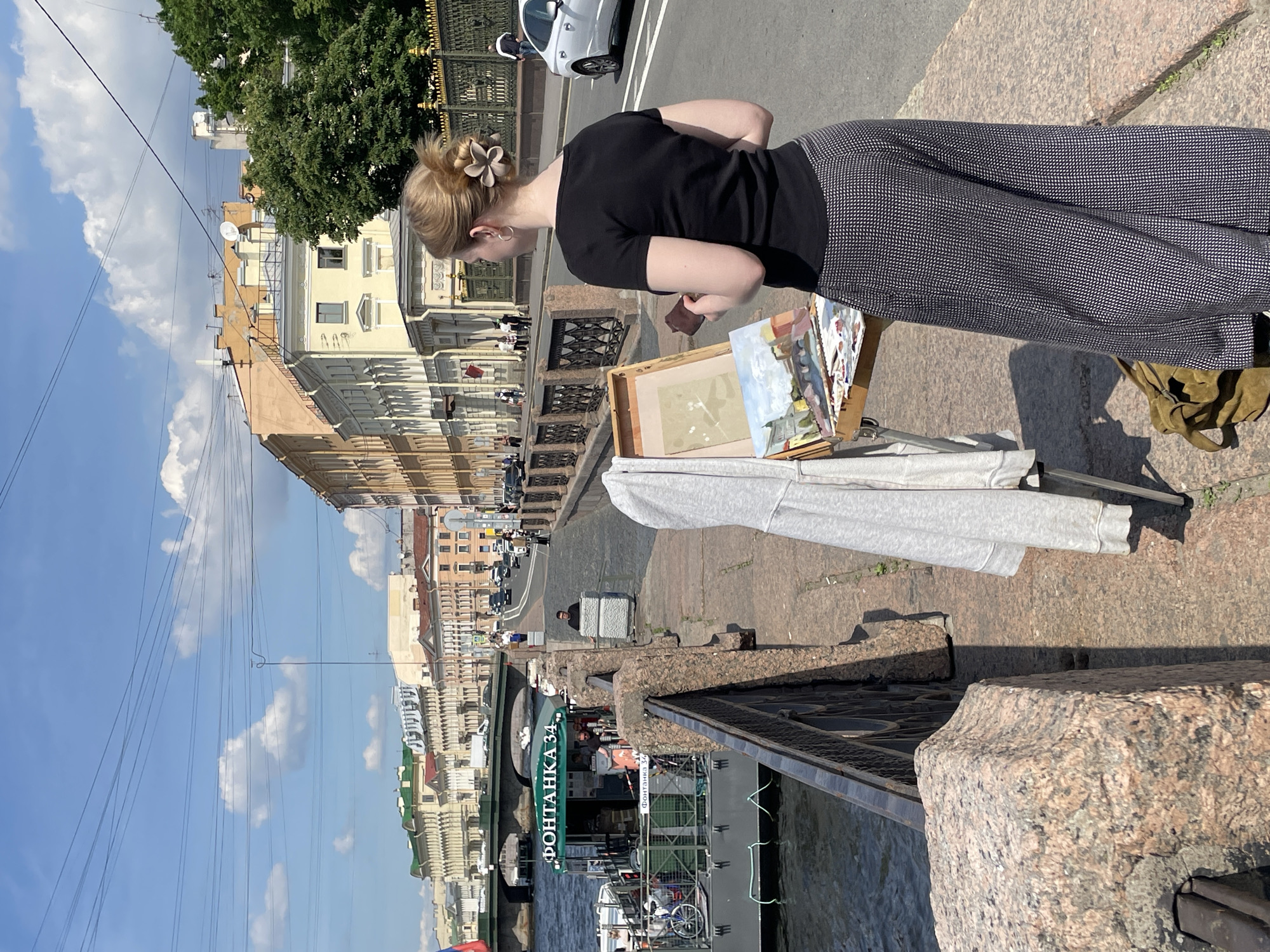 A young woman with blonde hair tied up with a flower accessory is painting on an easel on a sunny day. She is wearing a black t-shirt and wide-legged patterned pants. The easel holds a painting of a city landscape with buildings and a bridge. Various paint colors and brushes are on the easel's tray. A gray hoodie is draped over the easel. A brown bag is on the ground near the easel.
The scene appears to be along a canal in Saint Petersburg, Russia, as indicated by the sign on the boat which reads "ФОНТАНКА 34" (Fontanka 34). The boat is docked on the left side of the image and has people on board. In the background, there are numerous buildings with traditional European architecture, a bridge spanning the canal, and cars on a street. The sky is blue with scattered white clouds. The embankment is made of granite blocks and the street is paved with cobblestones.