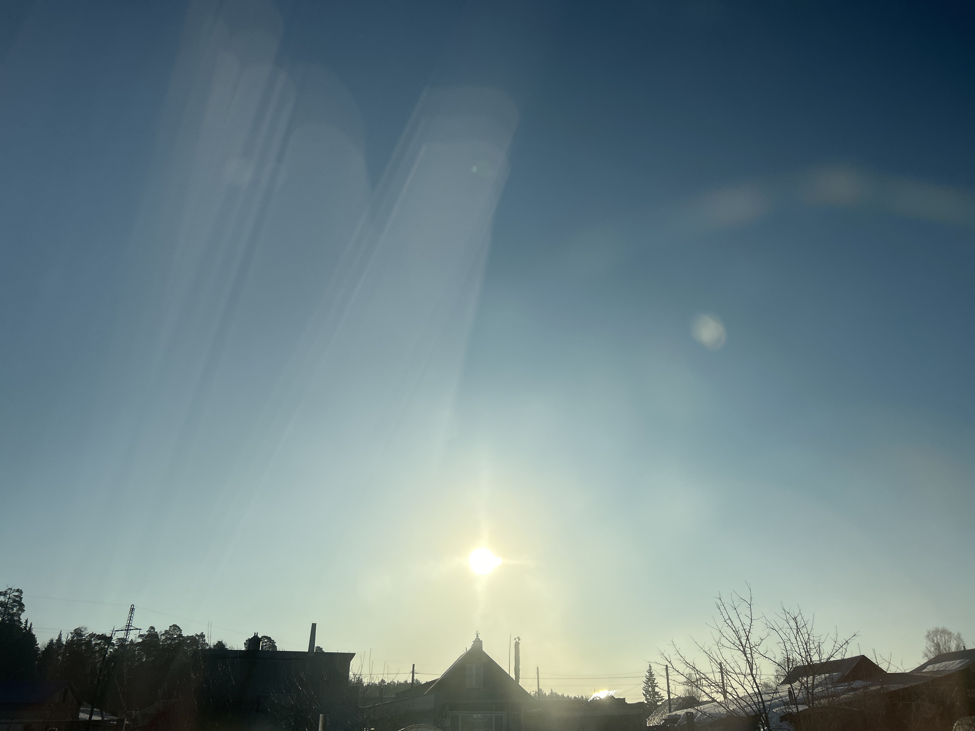 The image depicts a residential or suburban landscape in the city of Chaikovsky, Russia, observed during daylight hours under a clear, light blue sky. A bright, intense sun, positioned centrally in the lower third of the sky, produces significant lens flare and vertical sunbeams, backlighting the scene.
In the foreground and midground, a collection of buildings and natural elements are visible in silhouette. A prominent, light-colored gabled house with a dark roof and a central dormer window stands near the center of the lower frame, directly below the sun. To its left, a dark silhouette of a multi-tiered electricity pylon or large utility pole is visible above a dense line of coniferous trees. Several smaller residential structures with dark roofs, some partially covered in snow, are discernible to the left and right of the central house. Bare deciduous trees and sparse winter vegetation are present across the lower right portion of the frame, with some snow patches visible on the ground or roofs. Utility lines traverse the sky horizontally, particularly above the central house. No individuals or discernible actions are present in the scene.