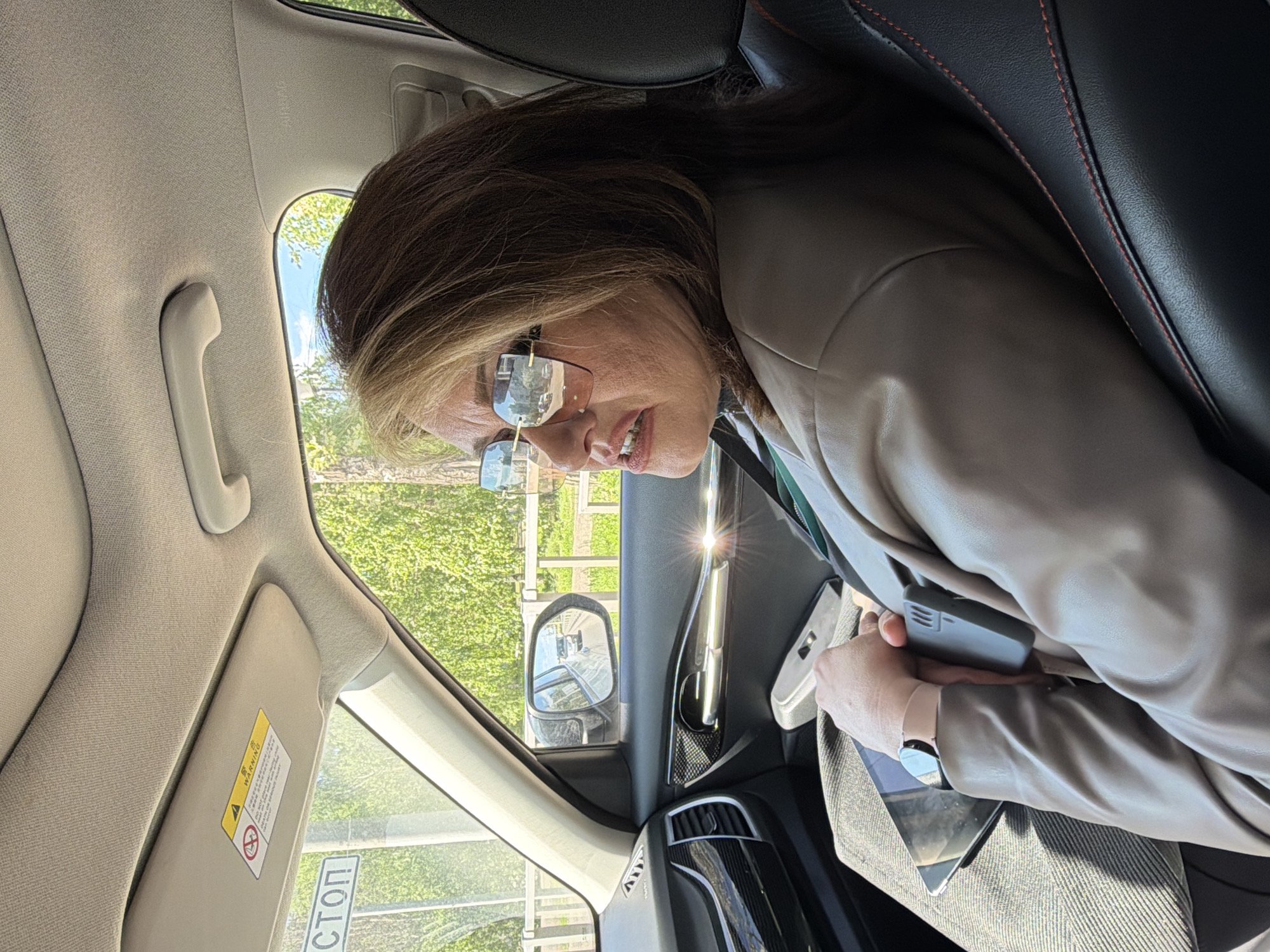 The image shows a woman inside a car. She is wearing rimless glasses and a light brown jacket. She is holding a phone and a tablet on her lap. A side mirror is visible, and a Russian "СТОП" sign is visible outside the car window. Based on the sign, the location is likely in Russia, though specific location within Krasnoyarsk cannot be determined from the information provided.