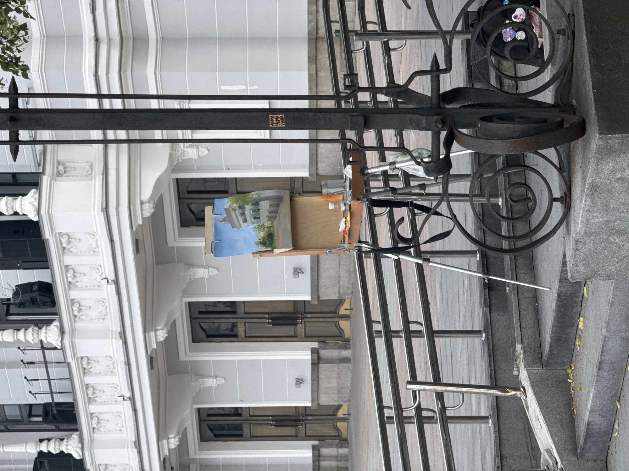 An outdoor scene depicts an easel with a painting of an old building set against a light gray building with ornate white trim and large dark doors. The easel holds art supplies, including bottles and brushes. A metal lamp post stands in the foreground, adorned with a small sticker. In the background, a metallic railing system leads up a ramp and stairs. A black backpack with heart-shaped pins is visible near the base of the lamp post. The overall setting suggests a public area in Yekaterinburg, Russia, likely a plaza or square near a historical building.