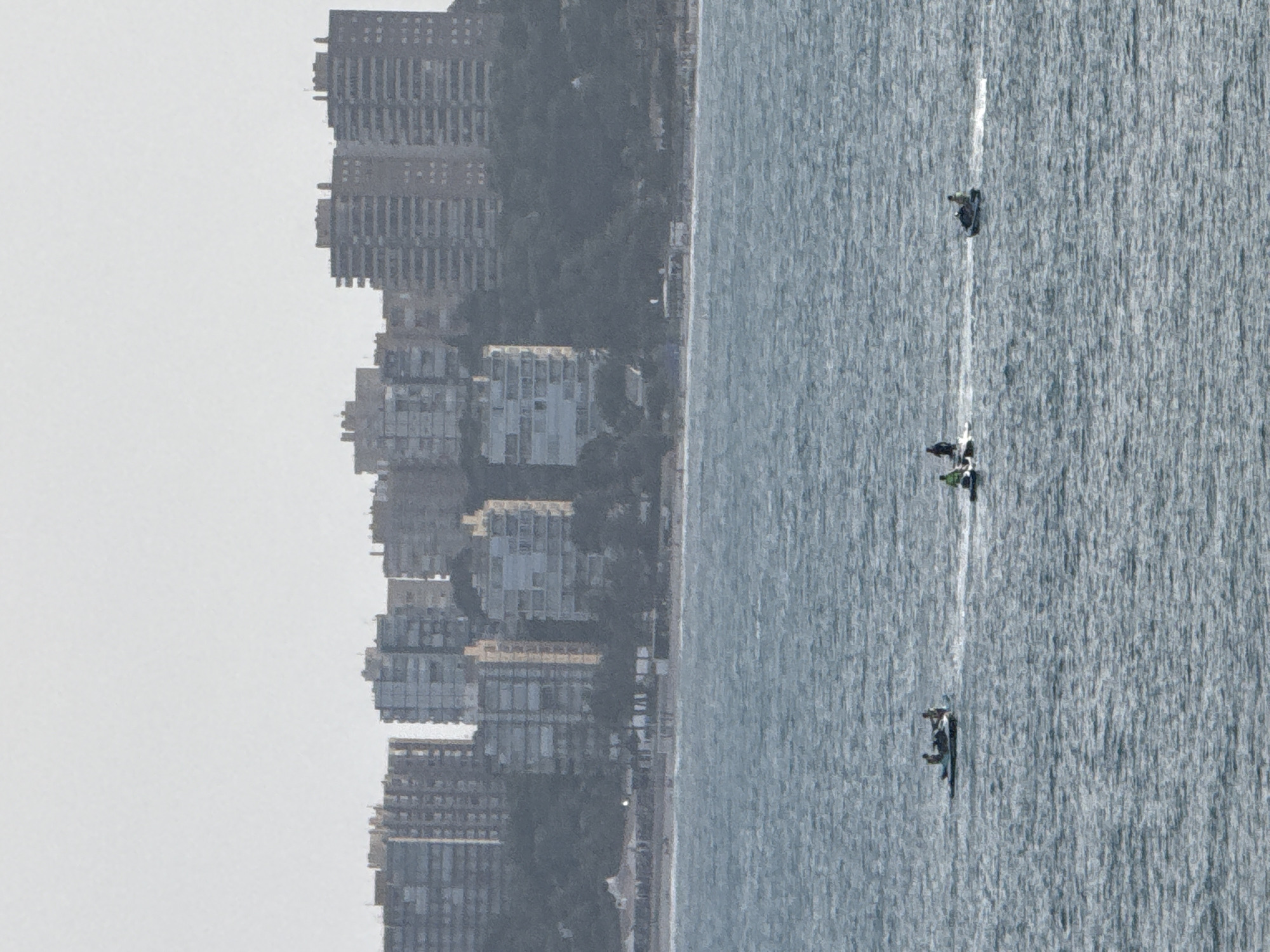 The image shows a wide view of the sea with a city skyline in the background. The sea is a choppy, deep blue. Three jet skis are visible on the water, each with at least one person riding it. The jet skis are spaced out across the frame. In the background, a dense collection of tall apartment buildings forms the city skyline. The buildings are mostly grey and white, with many windows visible. A thin strip of green trees and some beach can be seen between the city and the water. The sky is a pale, hazy white.
