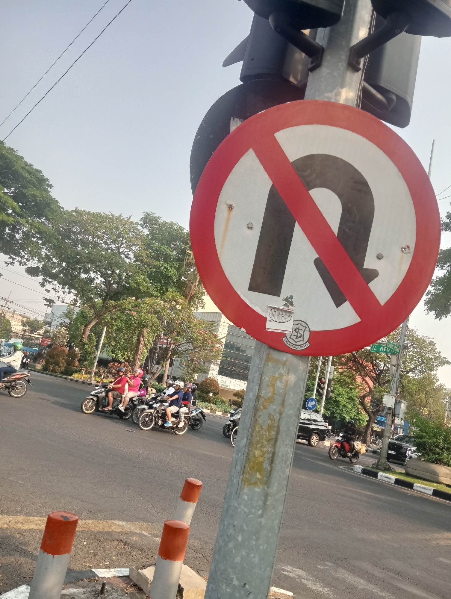 The image depicts a street scene in Surabaya, Indonesia, dominated by a prominent "no U-turn" traffic sign. The sign, a red circle with a diagonal red bar across a black U-shape on a white background, is mounted on a metal pole. Below the sign, a smaller sticker reads "DISHUB SURABAYA," indicating the city's transportation department. The street is paved and appears to be an intersection. Several people on motorcycles are visible, some riding in groups, navigating the road. A black car and a red motorcycle are also present. The environment is urban, with trees and modern buildings in the background. The sky is bright, suggesting daylight. The overall impression is of a busy city intersection with clear traffic regulations.