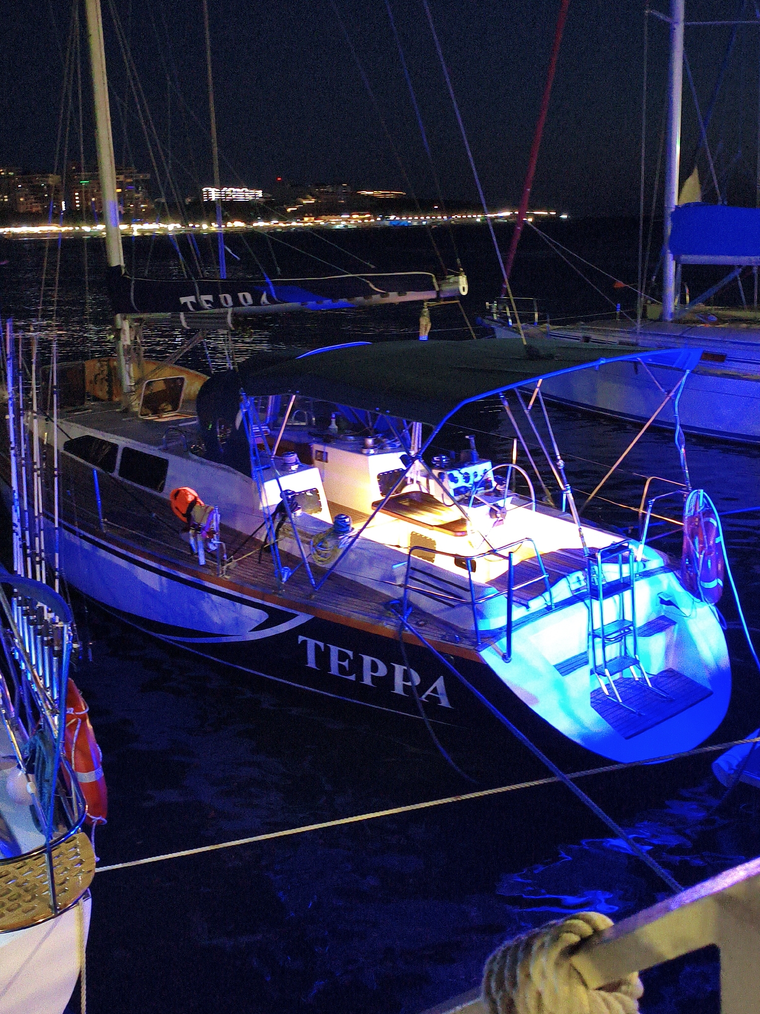 A nighttime view of moored yachts in the city of Krinitsa, Russia. The yachts are illuminated with blue and yellow lights, reflecting on the dark water. The name "TEPPA" is visible on the hull of one of the yachts. In the background, the city skyline is lit with numerous lights, suggesting a lively urban environment. The scene appears to be a serene harbor at night.
