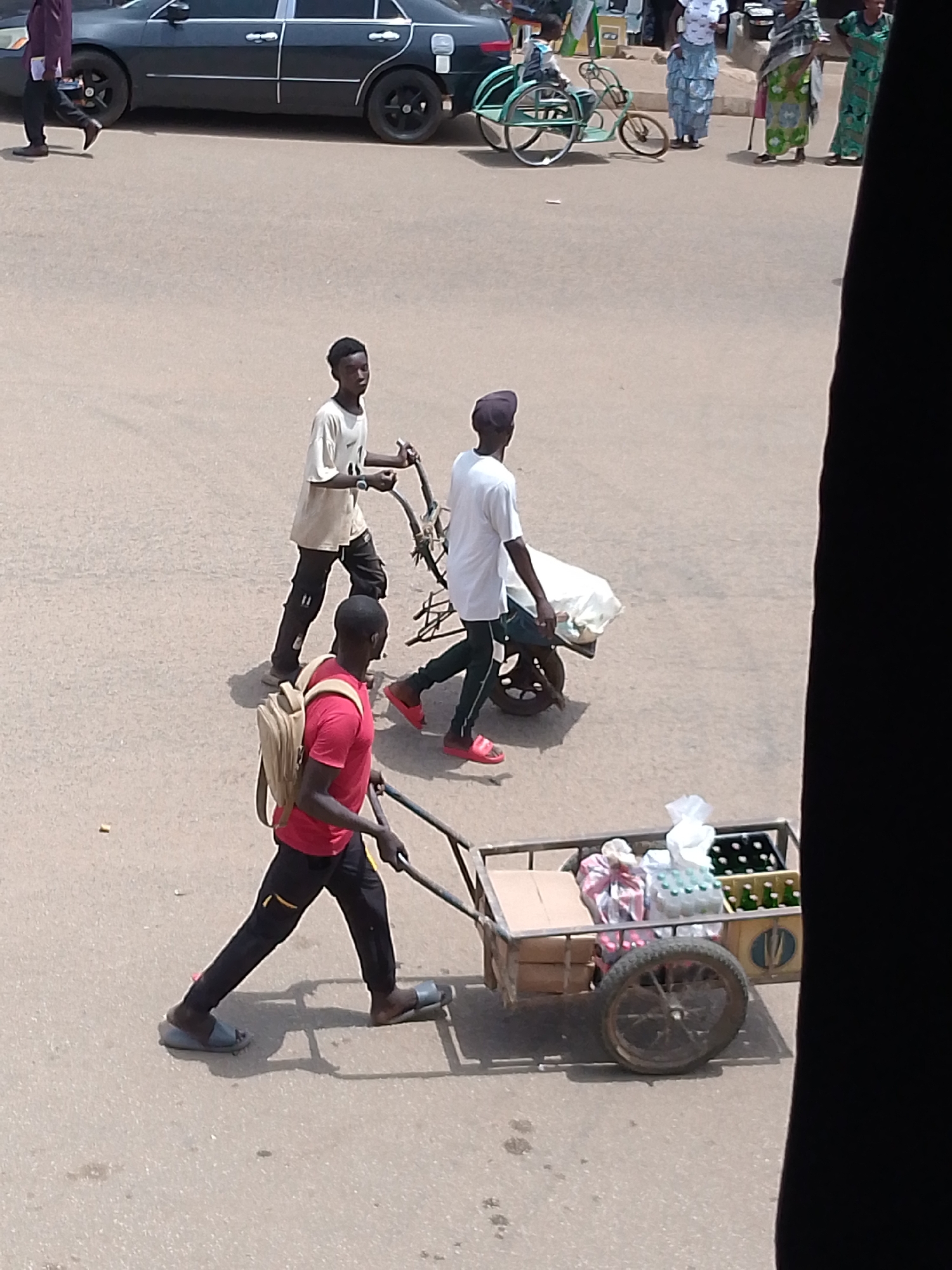 Three young men are seen on a paved street in Bukuru, Nigeria. The first man, wearing a red shirt, dark pants, a tan backpack, and blue sandals, pulls a metal cart containing bottled beverages and various items in cardboard boxes and plastic wrapping. The second man, clad in a white t-shirt, dark pants, and bare feet, pulls a small metal cart covered with a white cloth or bag. The third man, wearing a light-colored t-shirt and dark pants, is seen pulling what appears to be a metal structure. In the background, pedestrians and a dark-colored car are also visible. A disabled bicycle sits on the side of the street. The lighting suggests daytime.