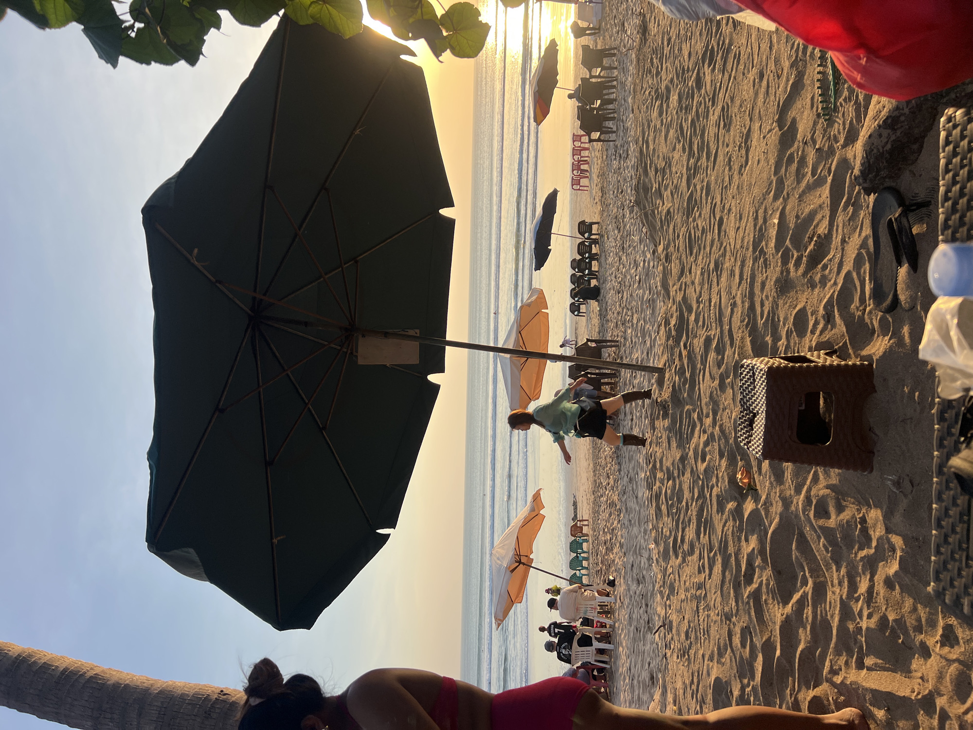 This image depicts a beach scene in Kuta, Indonesia, during sunset. The foreground is dominated by sand, with a small brown plastic stool in the center. To the left, a person's legs are visible, wearing pink shorts. In the middle ground, several beach umbrellas are set up, some with accompanying chairs. People are scattered around, some sitting at tables and others standing or walking. A woman in a green top and black shorts is captured in motion, appearing to be walking or dancing. In the background, the ocean stretches out to the horizon, with gentle waves rolling in. The sky is a warm gradient of orange and yellow, indicating the setting sun, which casts a golden glow over the entire scene. A palm tree trunk is visible on the far left, and some greenery hangs from the top right corner.