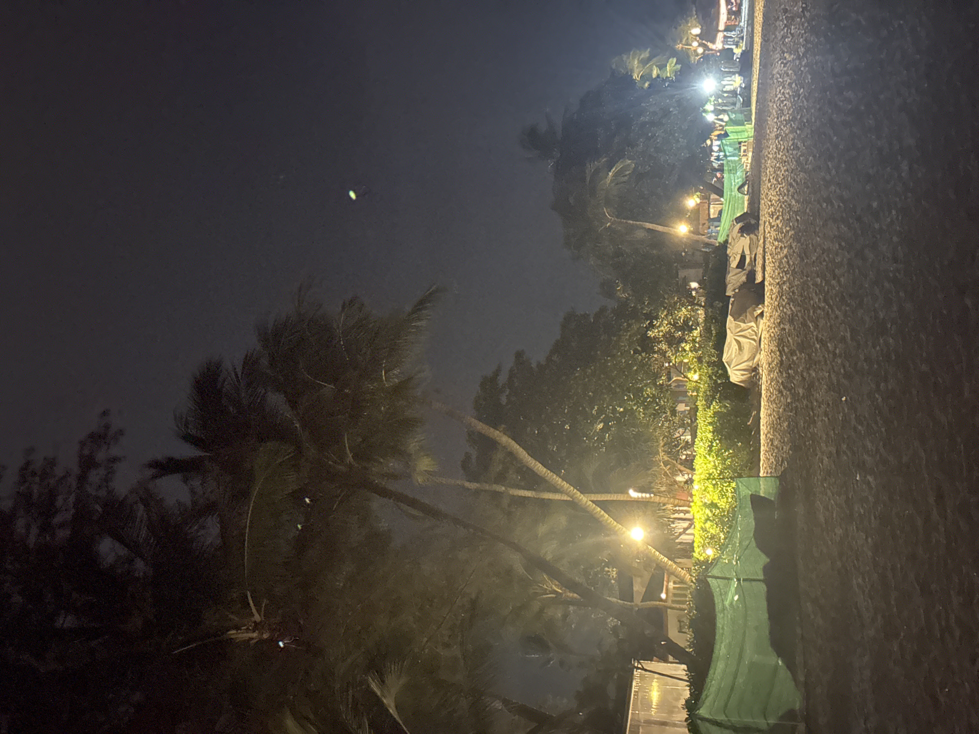 The image depicts a beach scene at night, likely in the city of Ko Samui District, Thailand, given the presence of palm trees and beach atmosphere. A dark sandy beach occupies the foreground. In the mid-ground, there is lush green foliage illuminated by artificial lights, with several palm trees silhouetted against the night sky. A temporary green barrier or tarp is erected on the left side of the frame. In the background, there are more trees, some structures, and a number of people gathered, suggesting a lively area or event. Several bright lights are visible, possibly from stalls, lamps, or vehicles, illuminating parts of the scene and creating a festive ambiance. The sky is dark, with a faint glow from artificial lighting. There is a blurry, light-colored object in the upper right quadrant of the sky, which could be a distant light or a lens artifact.