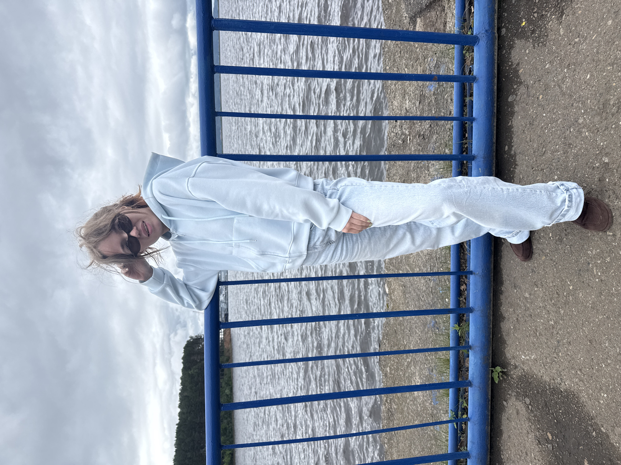 A young person is positioned slightly off-center to the right, standing on a paved surface beside a prominent blue metal railing, with a large body of water in the background. The individual, appearing to be female, wears dark sunglasses and has light brown, somewhat disheveled hair. She is dressed in a light blue, zip-up hooded sweatshirt and light-wash, wide-leg jeans, with dark brown flat shoes. Her left arm is resting on the top bar of the railing, and her left hand is raised towards her temple, touching her hair. Her right hand rests at her side.
The blue railing consists of a horizontal top bar, a lower horizontal bar, and multiple vertical balusters. Beyond the railing, the water surface is choppy and dark, reflecting an overcast sky overhead, which is heavily clouded. On the far bank of the water, a dense line of green trees is visible. A faint, light-colored vessel is discernible on the water in the distance. The ground beneath the subject is a dark gray, textured pavement (likely concrete or asphalt), with small patches of green vegetation growing near the base of the railing. The scene is located along a waterfront in the city of Chaikovsky, Russia.