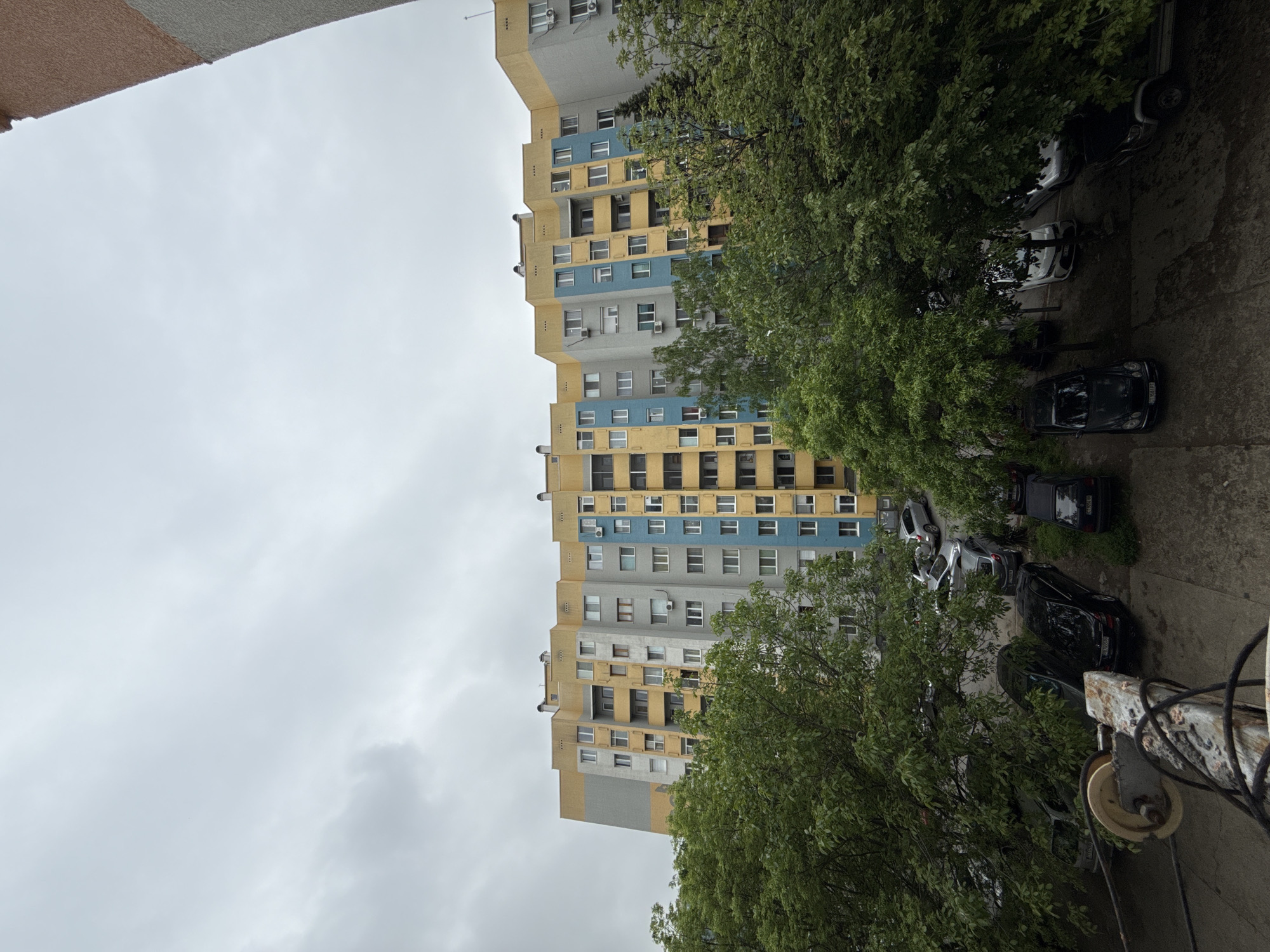 The image presents an upward view from what appears to be an apartment balcony in Stara Zagora, Bulgaria. The dominant subject is a tall, multi-story residential building, featuring a color scheme of yellow, blue, and gray. The building's facade has numerous windows, some with visible air conditioning units. In the foreground, lush green trees partially obscure the view. Beneath the trees, several parked cars are visible. The sky above is overcast, suggesting a cloudy or rainy day. Some miscellaneous electrical equipment is apparent in the bottom right corner of the image.